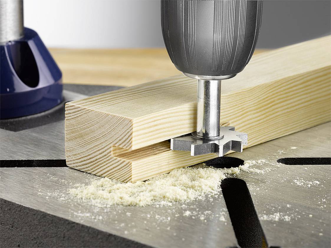 Een boor met een freesopzetstuk werkt aan een rechthoekig stuk hout op een werkbank, waarbij het hout nauwkeurig wordt bewerkt.