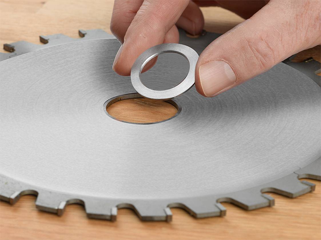 Een hand houdt een metalen ring vast boven een groot zaagblad op een houten tafel.