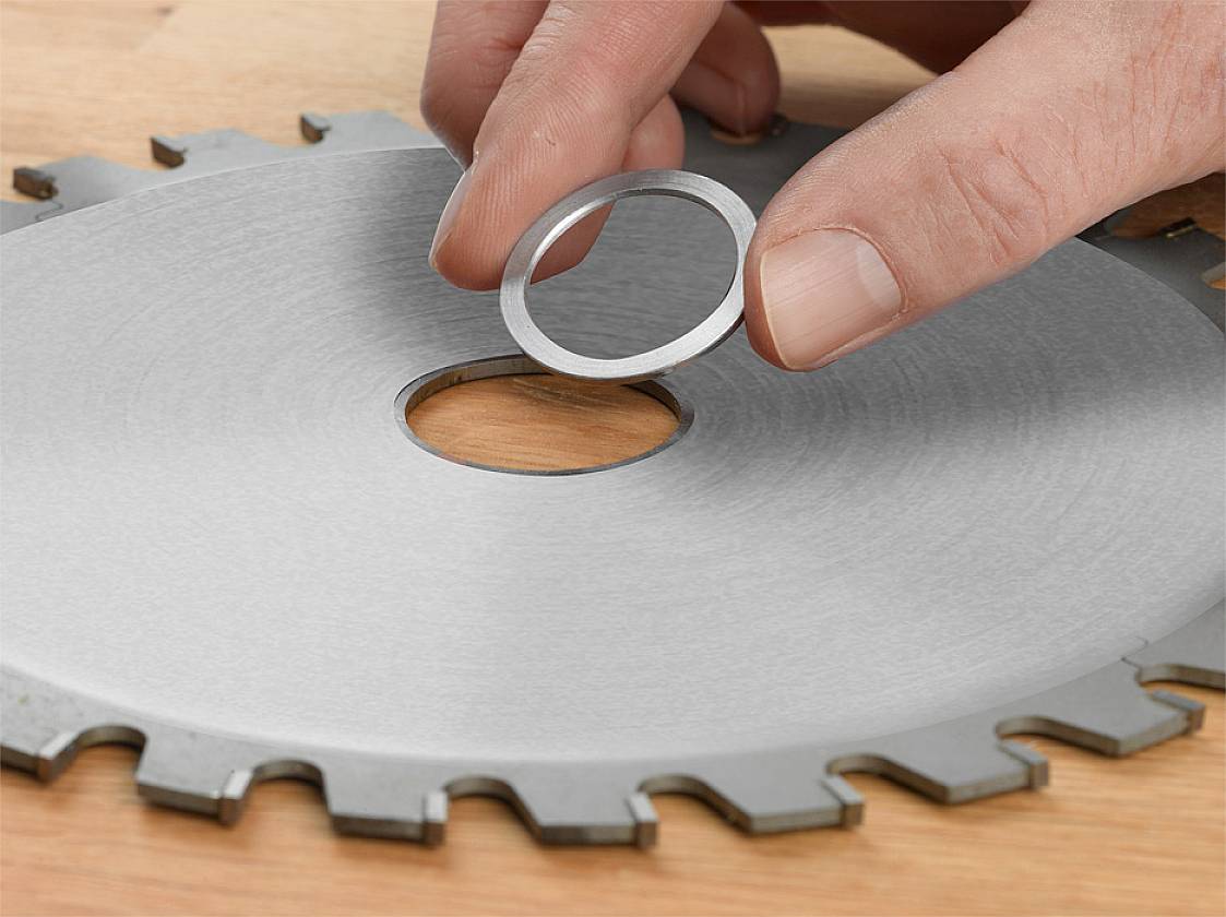 Een hand houdt een kleine metalen ring boven een ronde zaagblad-opening op een houten oppervlak.