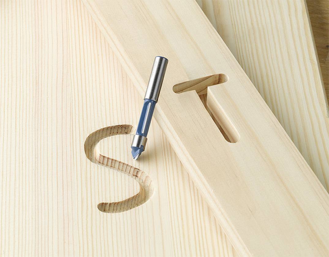 Een houten boor ligt naast een stuk hout met de letter 'S' erin gegraveerd. Ernaast bevindt zich een sleufvormig gat in het hout.