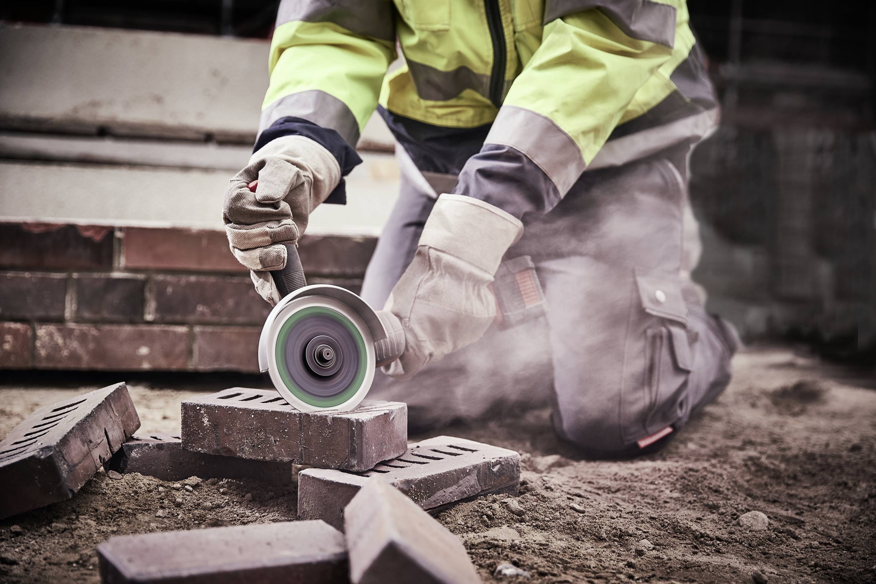 Een arbeider in beschermende kleding snijdt stenen met een slijpmachine op een bouwplaats, waarbij stof opwaait.