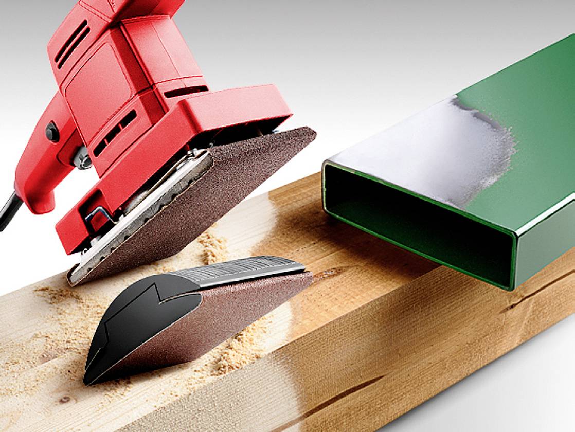 Een rode slijpmachine slijpt een houten plank. Ernaast ligt een groen, rechthoekig metalen buisje dat half geslepen is.