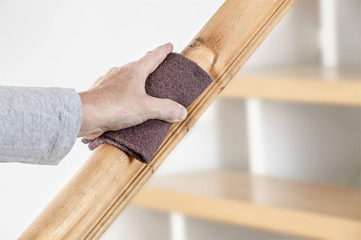 Een hand schuurt een houten leuning met schuurpapier. De beweging is cirkelvormig om het hout glad te maken en voor te bereiden.