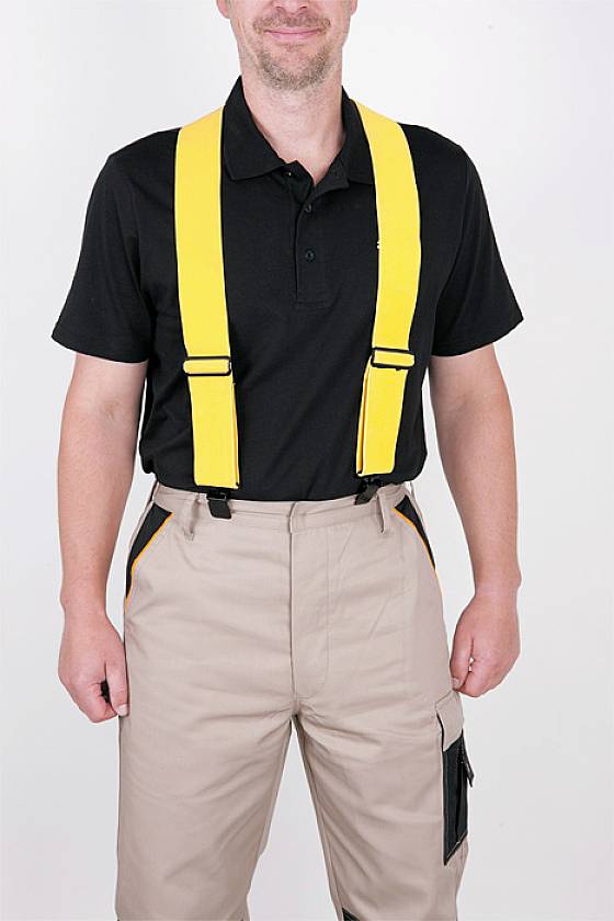 Een man in een zwart poloshirt en beige broek met gele bretels staat voor een witte achtergrond.