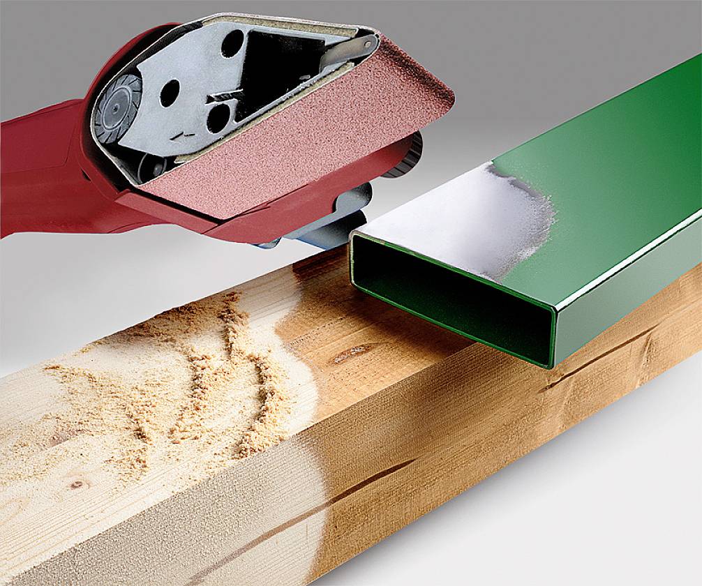 Een rode bandschuurmachine bewerkt een stuk hout dat onder een groene metalen drager ligt. Houtspaanders zijn zichtbaar op het houtoppervlak.