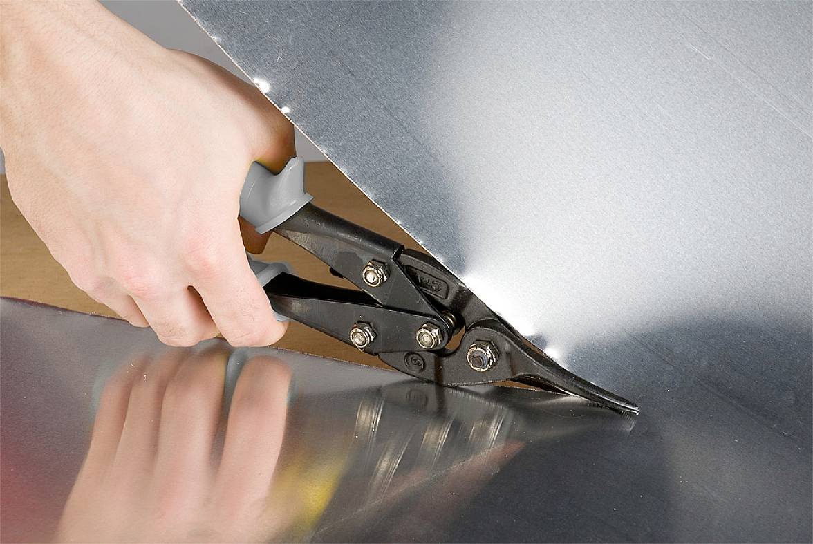 Een hand gebruikt een blikschaar om een metalen plaat te knippen. Het gereedschap heeft zwarte handgrepen en ligt op een gladde ondergrond.