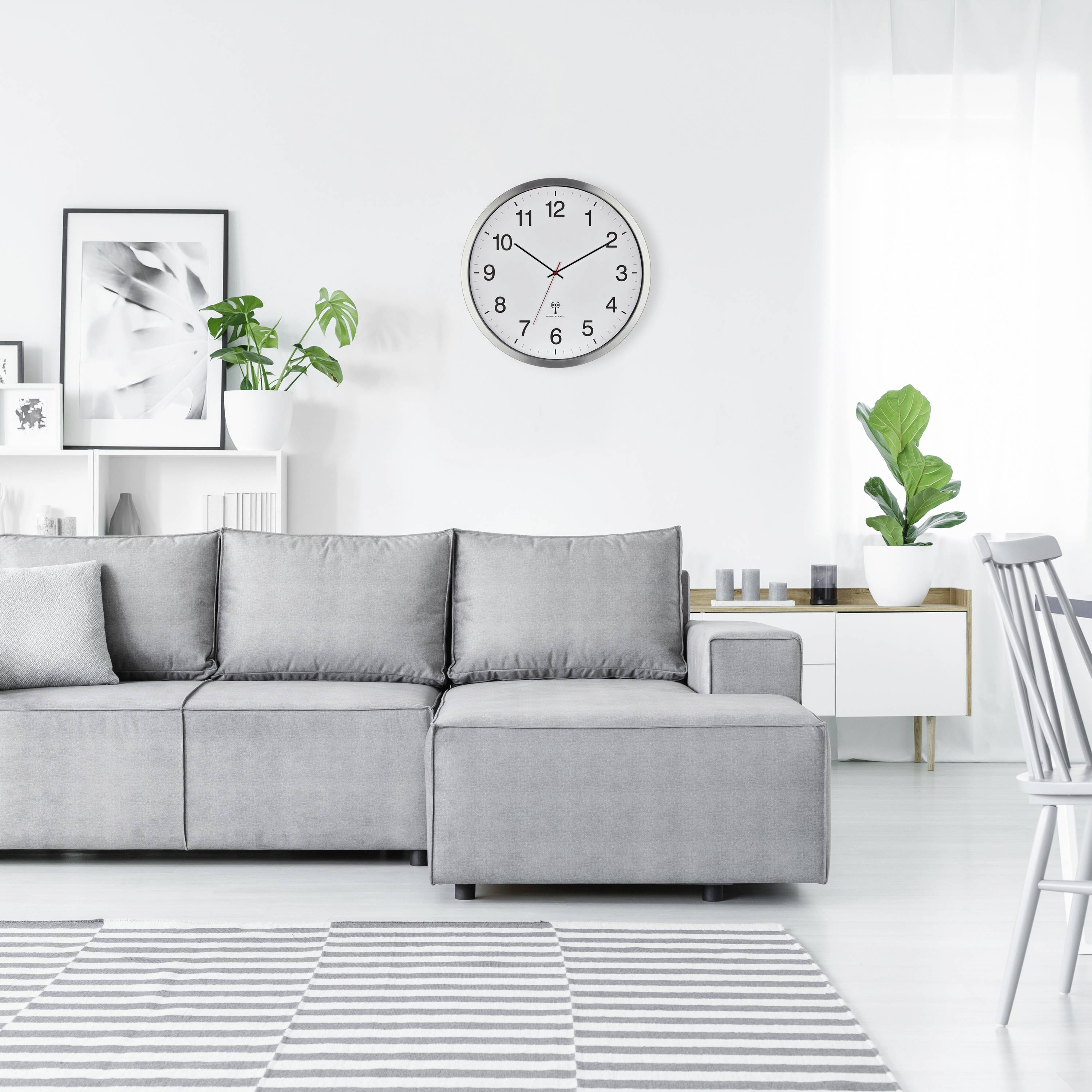 Grijs bankstel in een lichte woonkamer met wandplanken, planten en een ronde klok. Moderne, minimalistische interieurstijl.