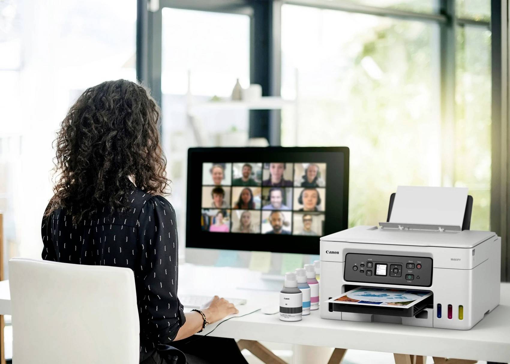 Een vrouw zit aan een bureau en neemt deel aan een videoconferentie op een computerscherm. Naast haar staat een printer.