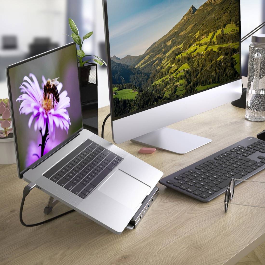 Een laptop met een bloemenbeeldscherm staat naast een grote desktopmonitor met een berglandschap op een bureau met toetsenbord en muis.