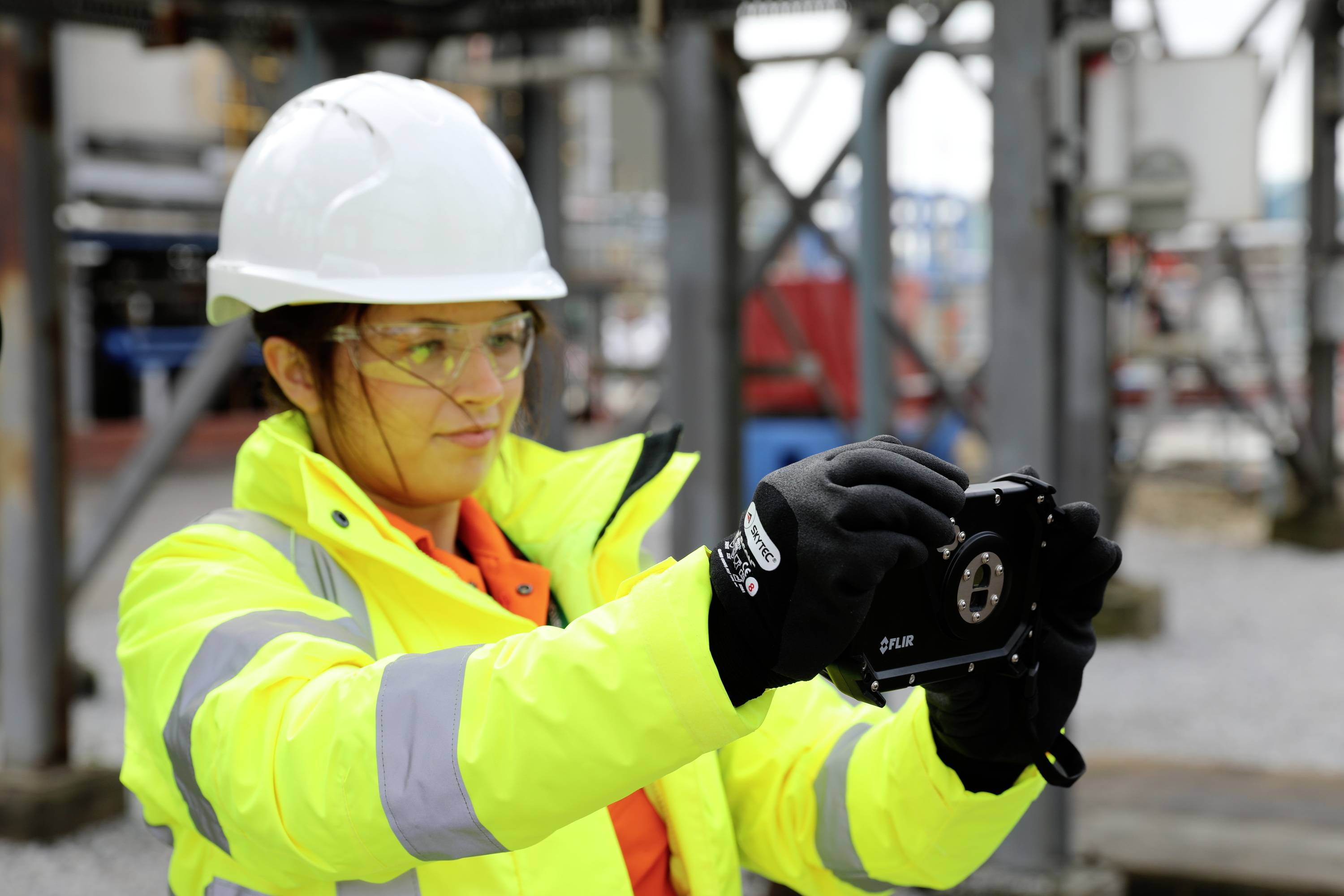 Een persoon in beschermende uitrusting, inclusief helm en veiligheidsbril, bedient een technisch apparaat op een industriële bouwplaats.