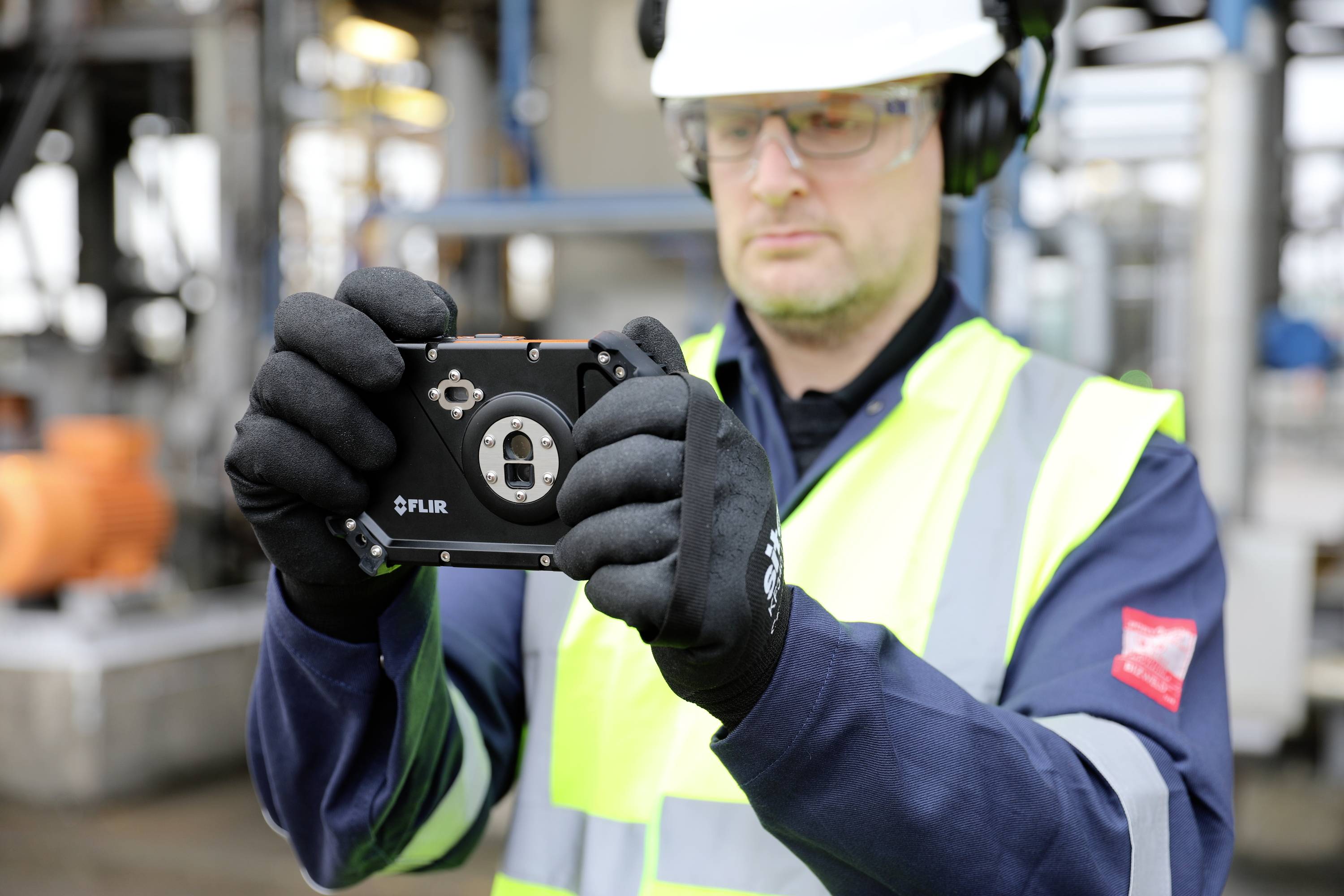 Een werknemer in beschermende kleding en helm houdt een meetinstrument vast, geconcentreerd aan het werk in een industriële omgeving.