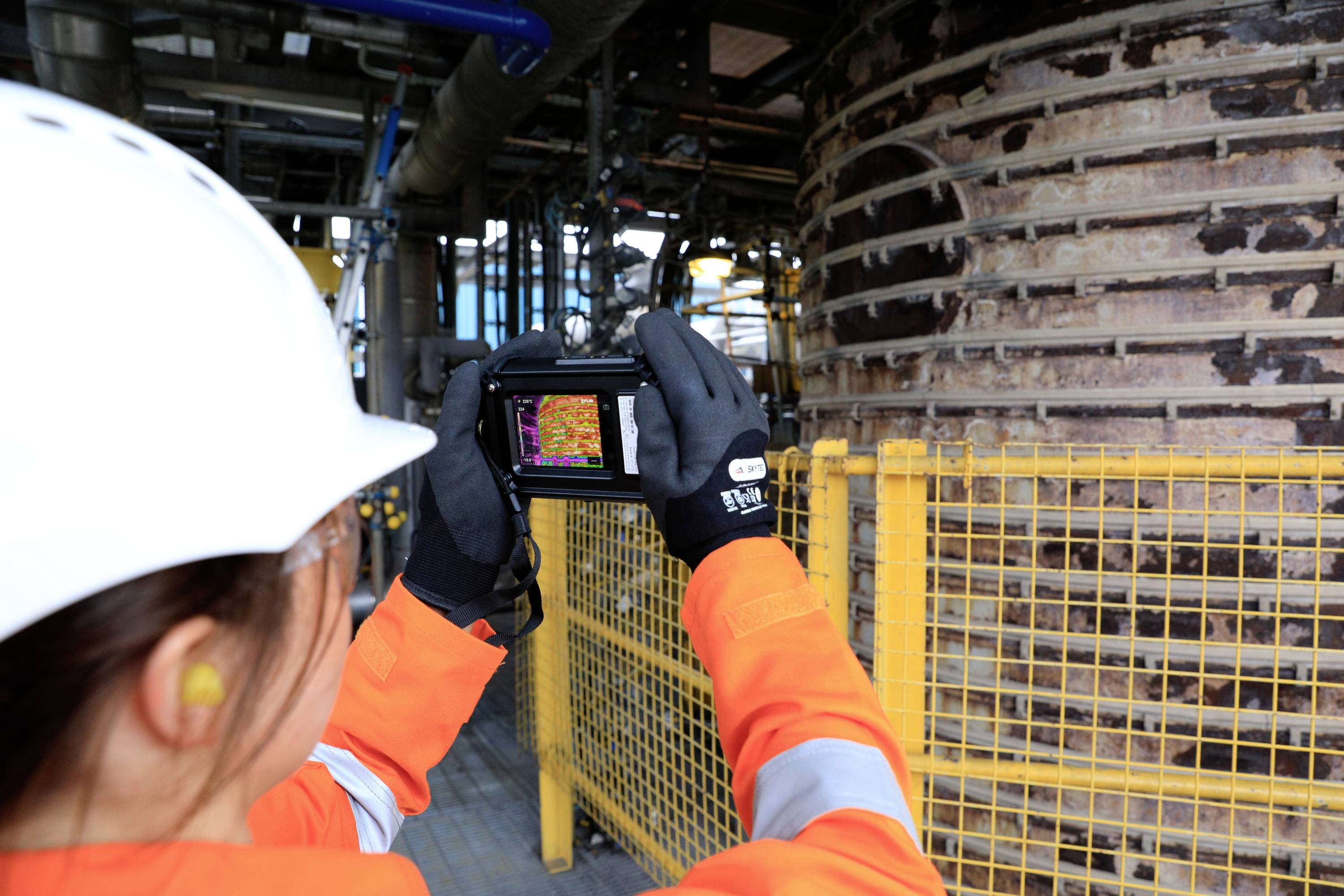 Een persoon in beschermende kleding fotografeert een industriële installatie met een warmtebeeldcamera. Een grote metaalstructuur is zichtbaar.
