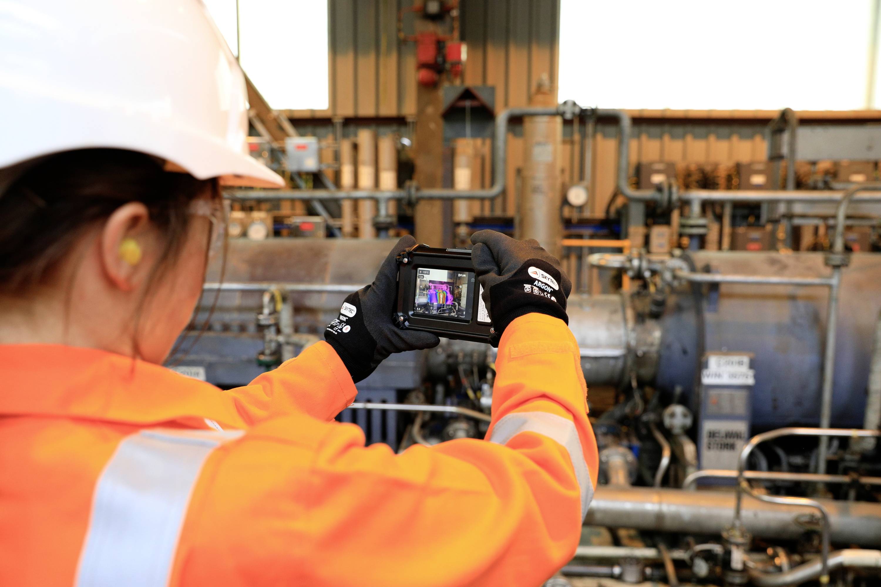 Een persoon in oranje beschermende kleding en helm gebruikt een camera-apparaat in een industriële omgeving met buizen en machines.