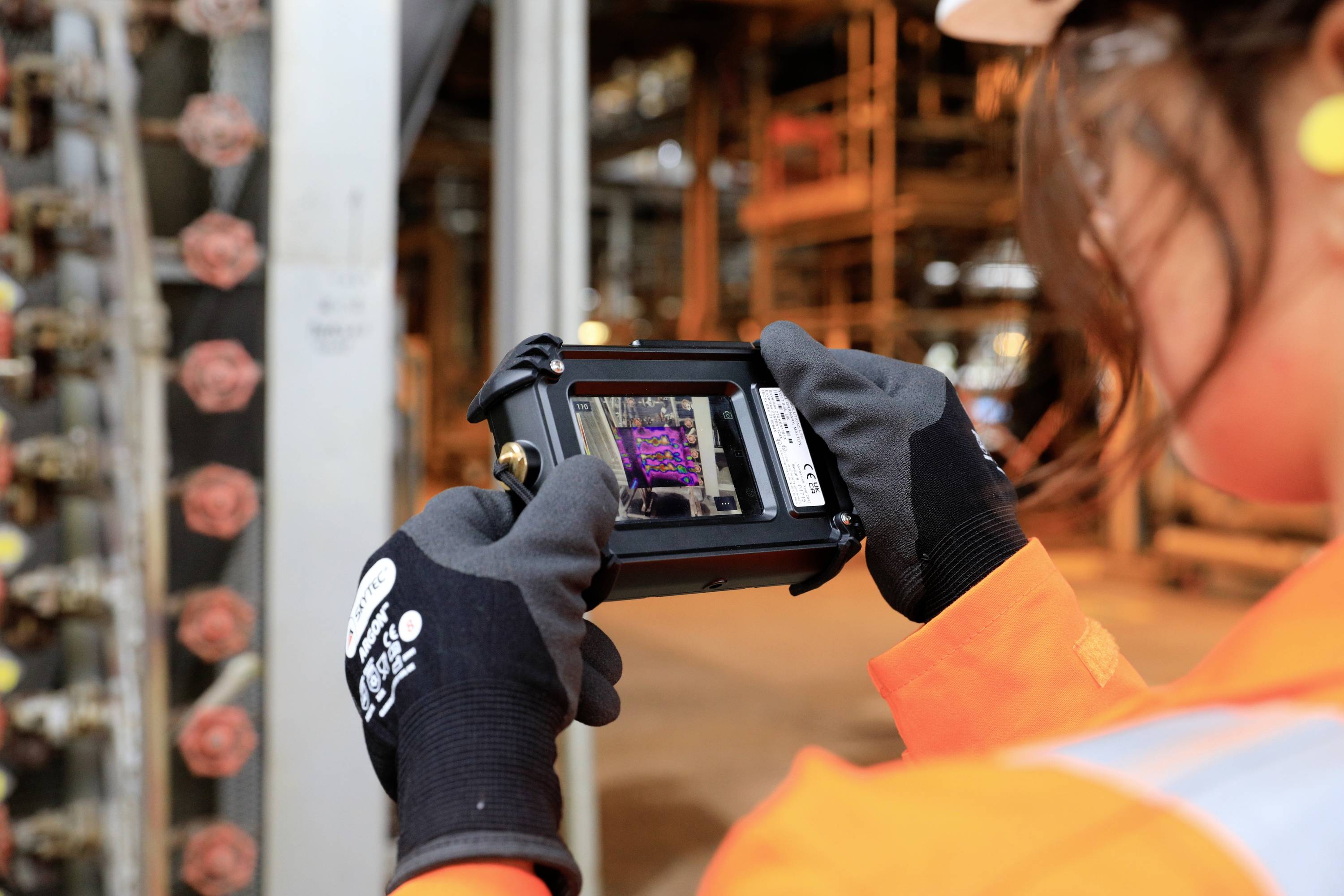 Een persoon in werkkleding en helm fotografeert met een draagbaar apparaat een industriële omgeving in een fabriekshall.