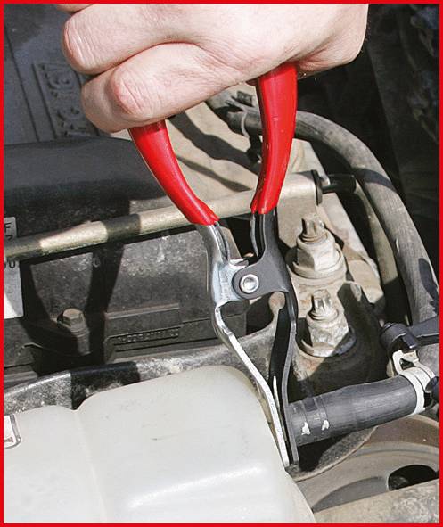 Een hand bedient een rode tang om een metalen voorwerp los te maken van een auto-onderdeel. De afbeelding toont mechanisch reparatiewerk.