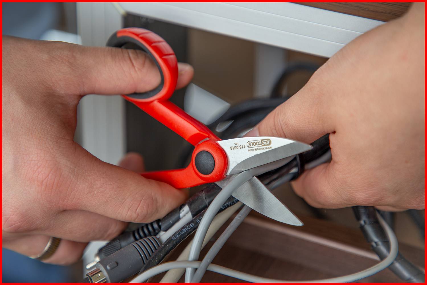 Twee handen houden een rode schaar vast en knippen verschillende zwarte en grijze kabels door. Links is een deel van een meubelstuk zichtbaar.