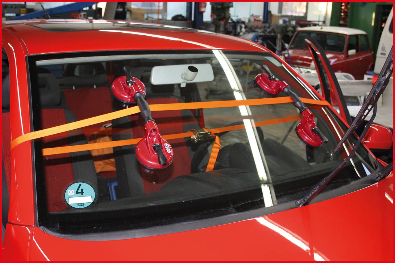 Rode auto in werkplaats met nieuw geplaatste voorruit. Vacuümzuigers borgen de ruit, oranje draaghulpstukken zorgen voor stabilisatie.