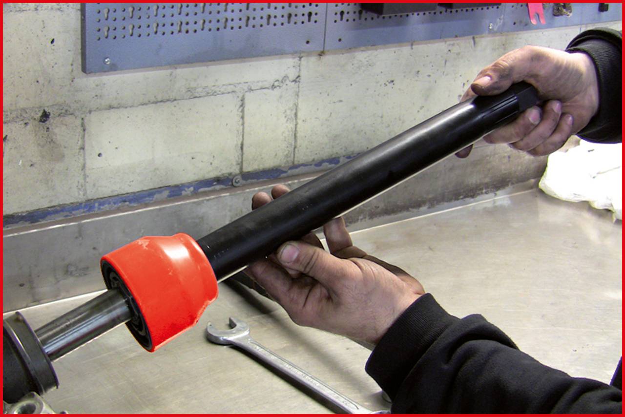 Een persoon houdt een zwart metalen pijp met een feloranje beschermkap in een werkplaats. Naast de pijp ligt een moersleutel.