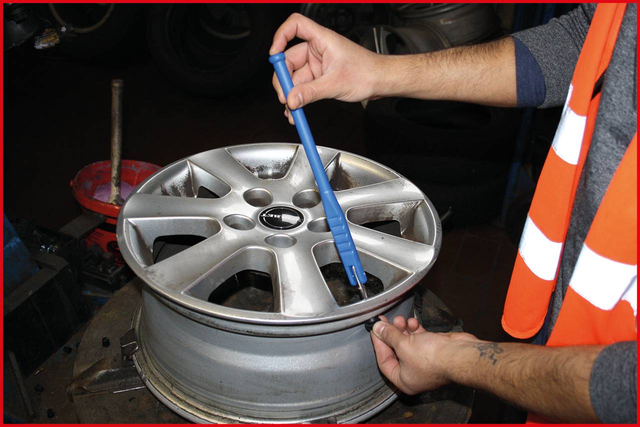 Een persoon controleert de velg van een auto met een blauw gereedschap. Hij draagt een oranje veiligheidshesje.