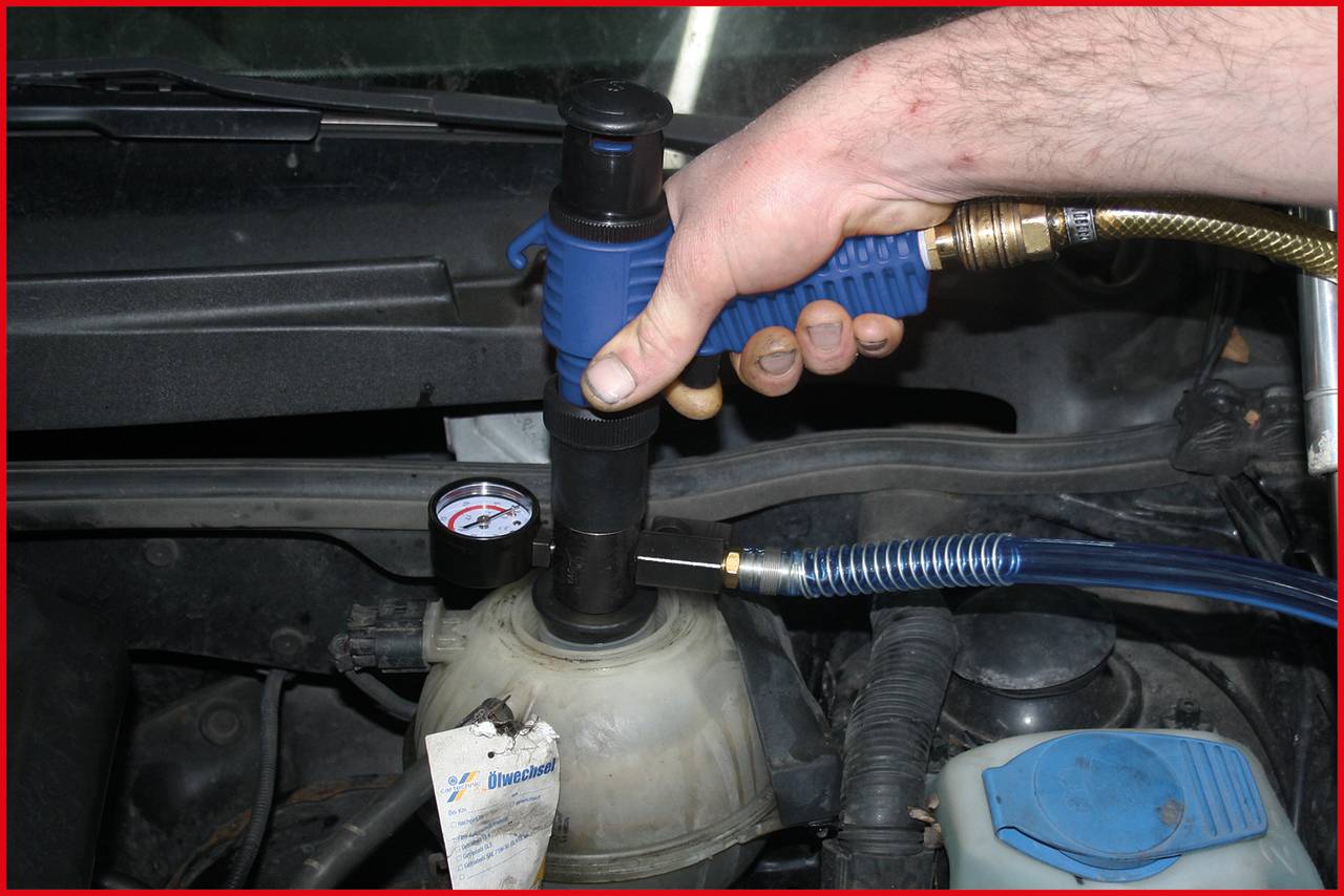 Een hand houdt een blauwe gereedschapspistool vast dat is aangesloten op het koelvloeistofreservoir van een auto, terwijl een manometer de druk aangeeft.