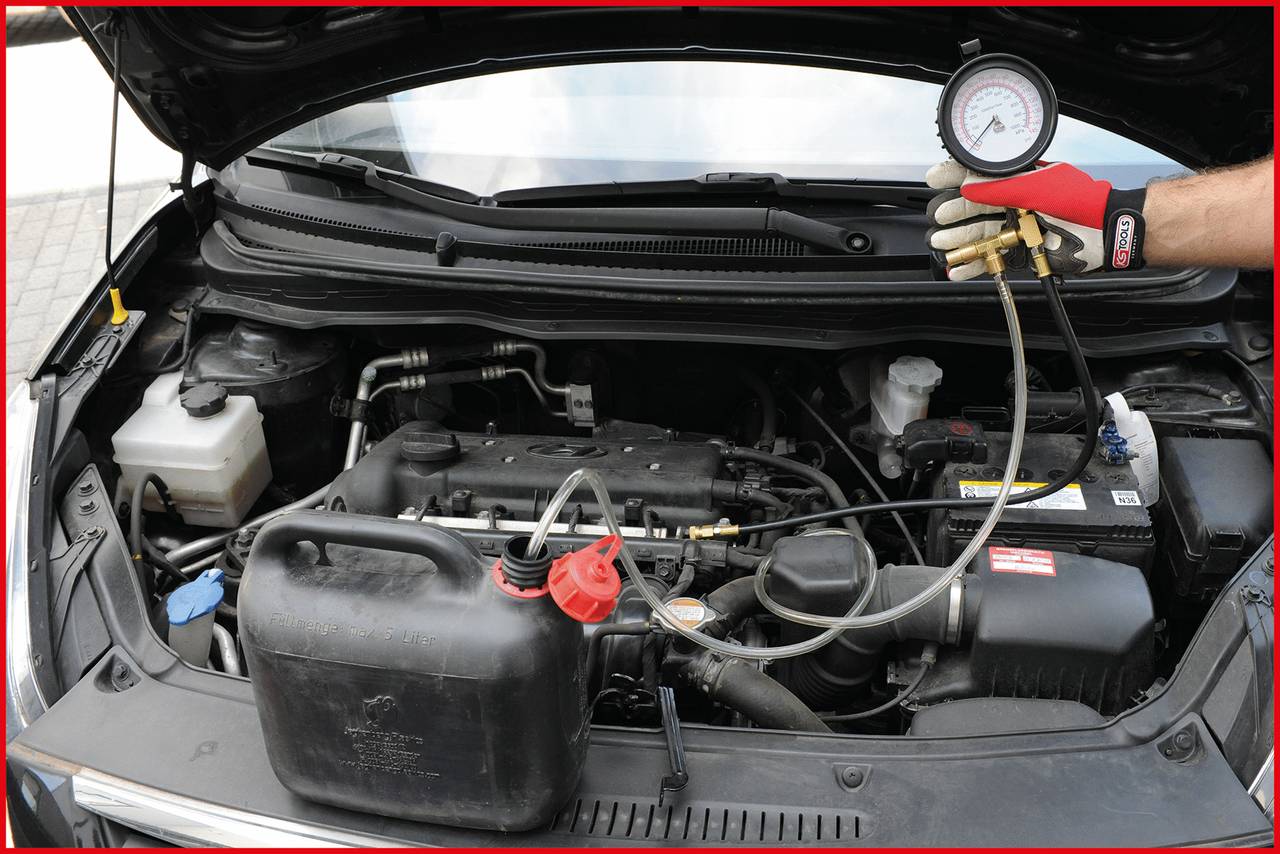 Een geopende automotorruimte, een monteur houdt een manometer vast om de druk te controleren. Op de voorgrond staat een zwarte jerrycan.