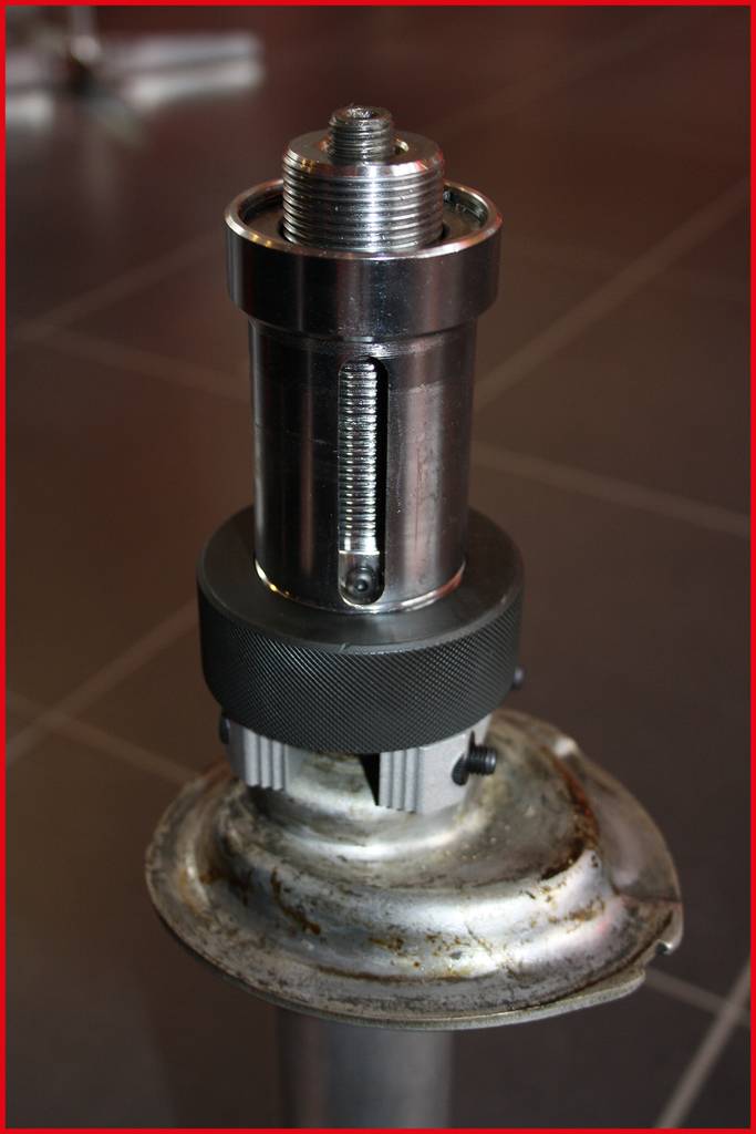 Een zilveren mechanisch apparaat met veerwerk op een metalen basis, geplaatst in een werkplaatsomgeving.