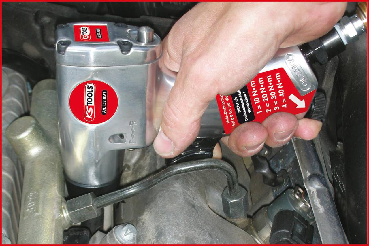 Een hand houdt een KS Tools pneumatische slagmoersleutel vast, die op een schroef in een motorruimte wordt gezet om deze los te draaien.