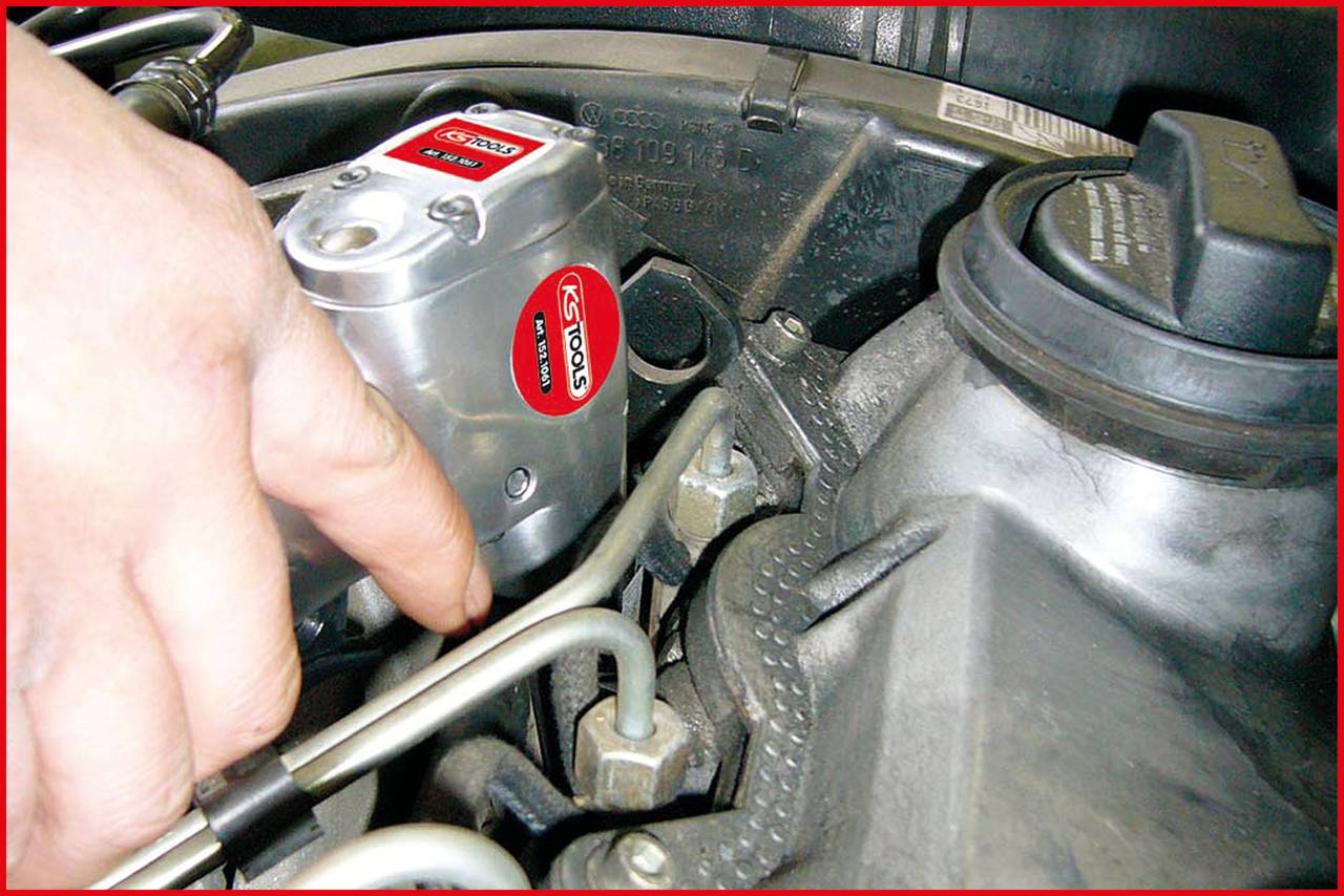 Een hand houdt een metalen Koso-onderdeel vast in de motorruimte van een auto. Kabels en slangen omringen het onderdeel.