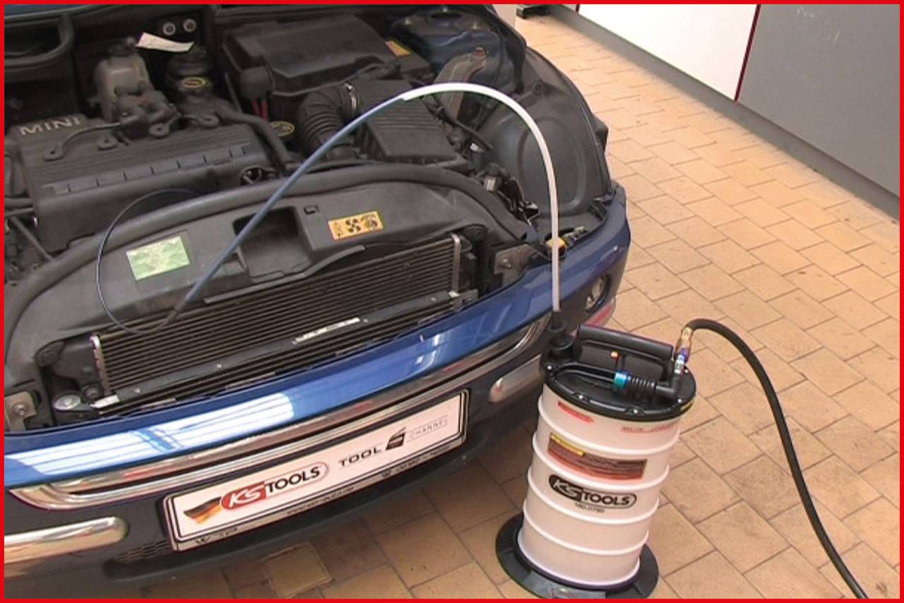 Een auto met geopende motorkap staat in een werkplaats. Een slang is verbonden met een apparaat dat olie aftapt.