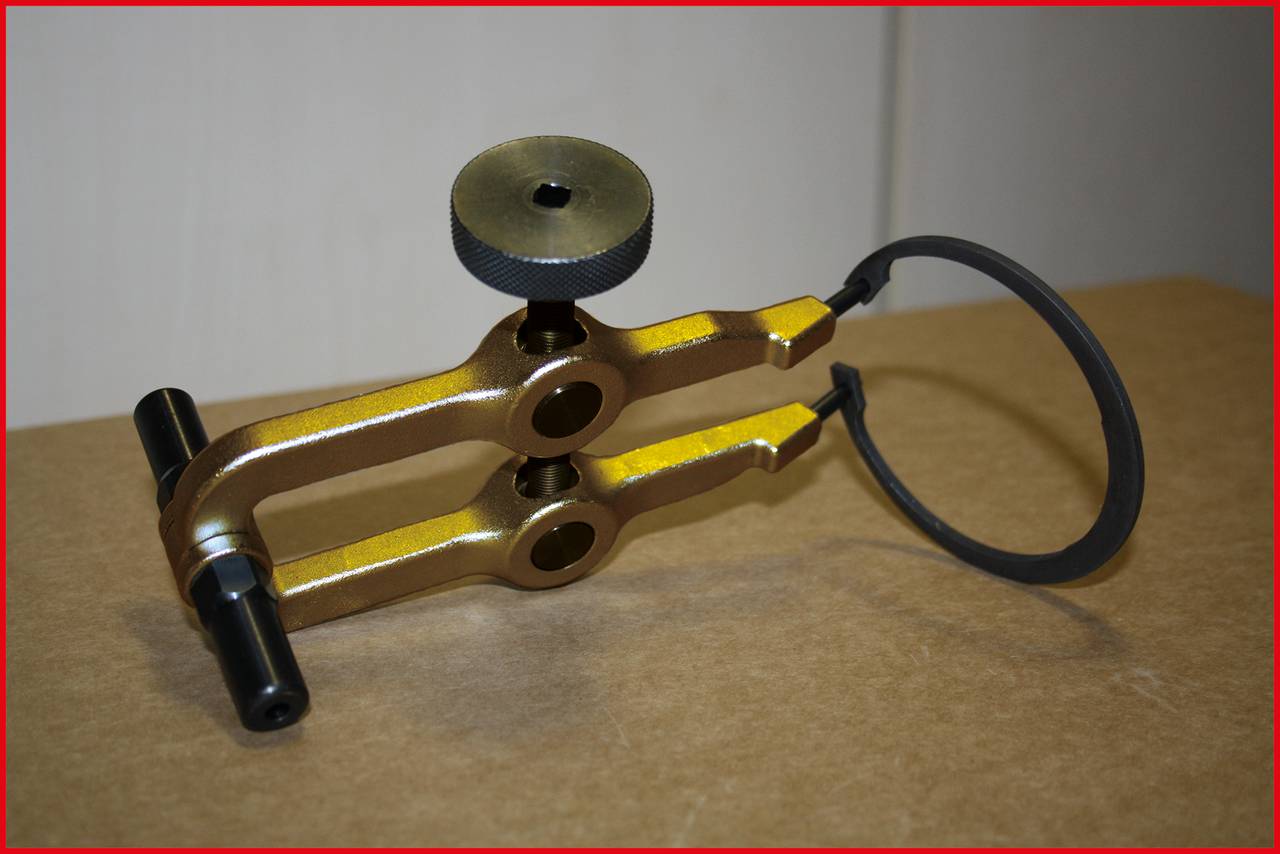 Metalen gereedschap geplaatst op een tafel, met een hefboomconstructie en een zwart wiel. Het lijkt bedoeld voor nauwkeurige mechanische werkzaamheden.