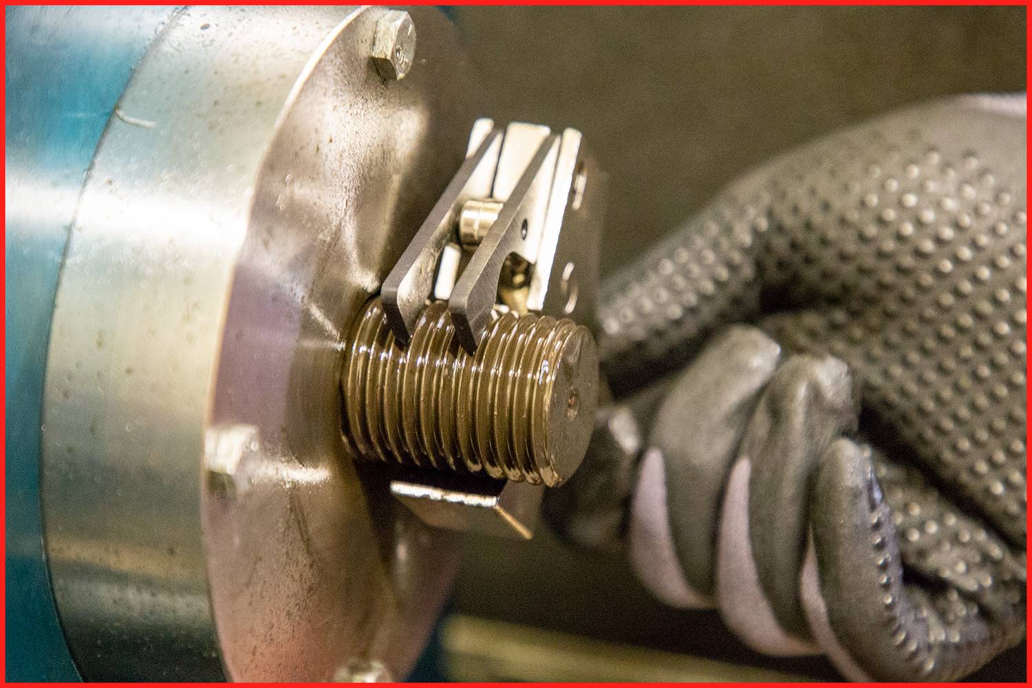 Een hand in een beschermende handschoen houdt een metalen gereedschap vast aan een cilindrische metalen machine, waarschijnlijk in een werkplaats.