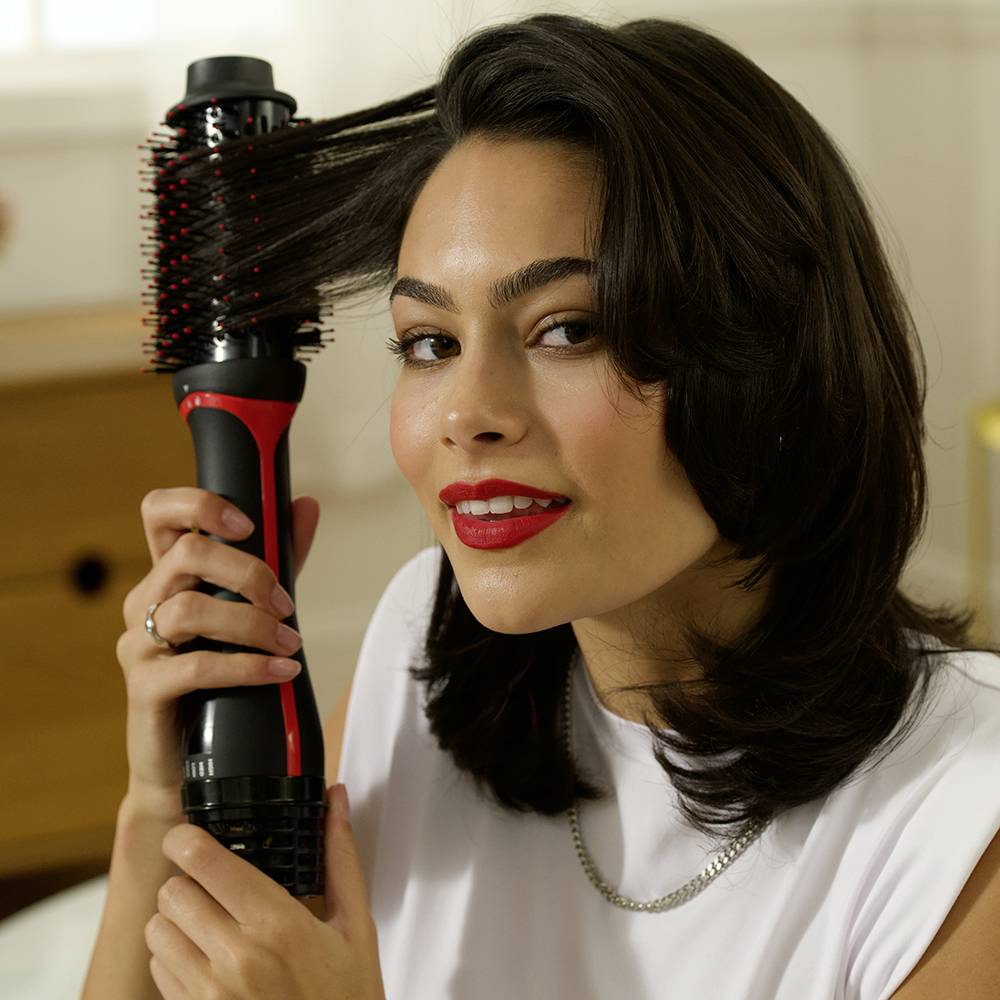 Een vrouw gebruikt een elektrische föhnborstel om haar haar te stylen. Ze glimlacht licht naar de camera, haar haar is glad en glanzend.