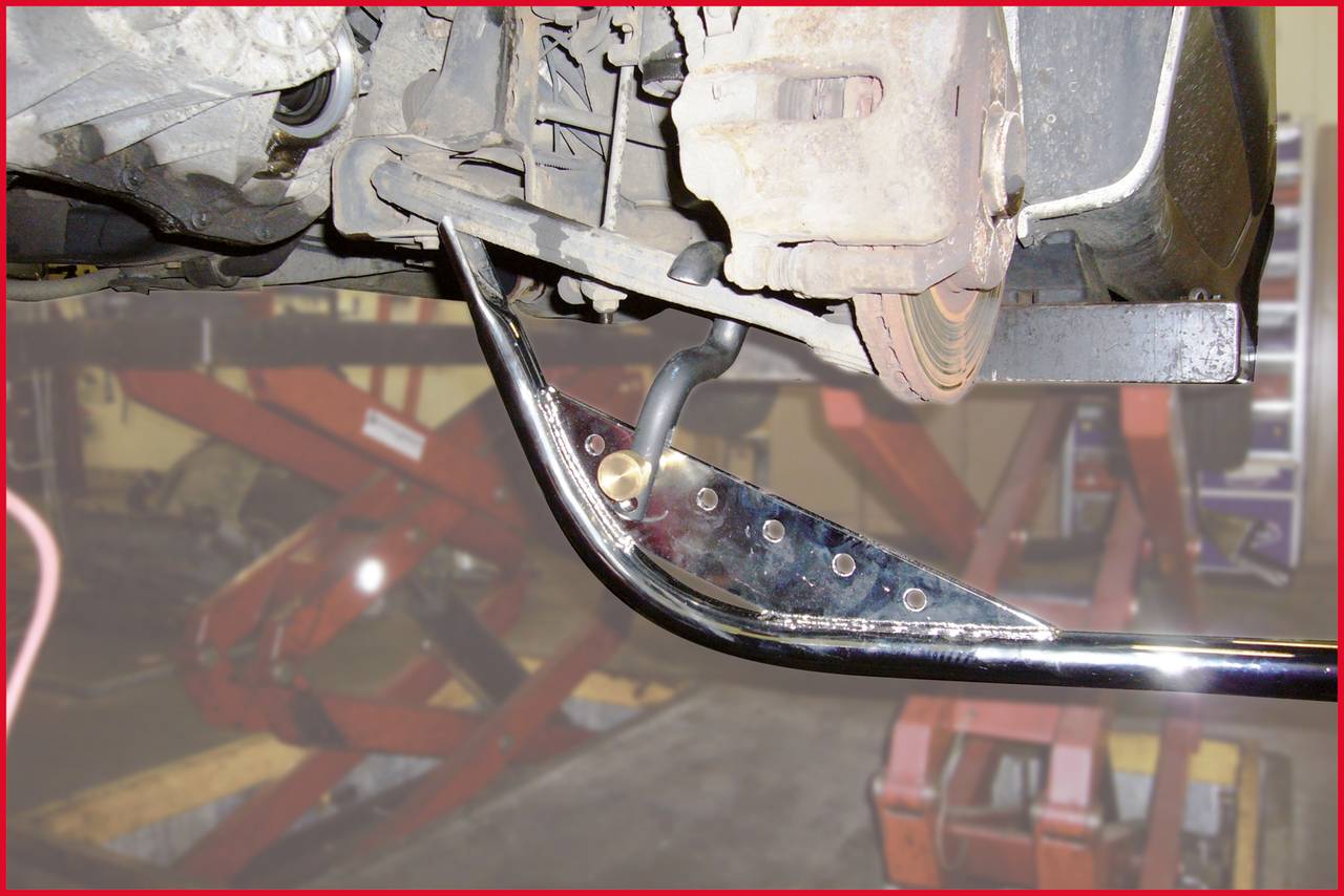 Een auto staat op een hefbrug, een metalen onderdeel wordt gerepareerd of gemonteerd. Op de achtergrond zijn autoreparatiegereedschappen zichtbaar.
