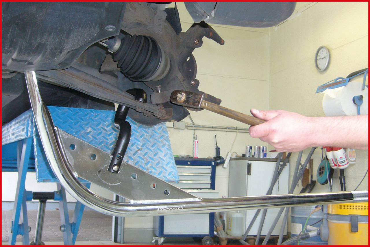 Een persoon gebruikt een gereedschap om aan de achterste wielophanging van een auto te werken. Op de achtergrond zijn werkplaatsgereedschappen zichtbaar.