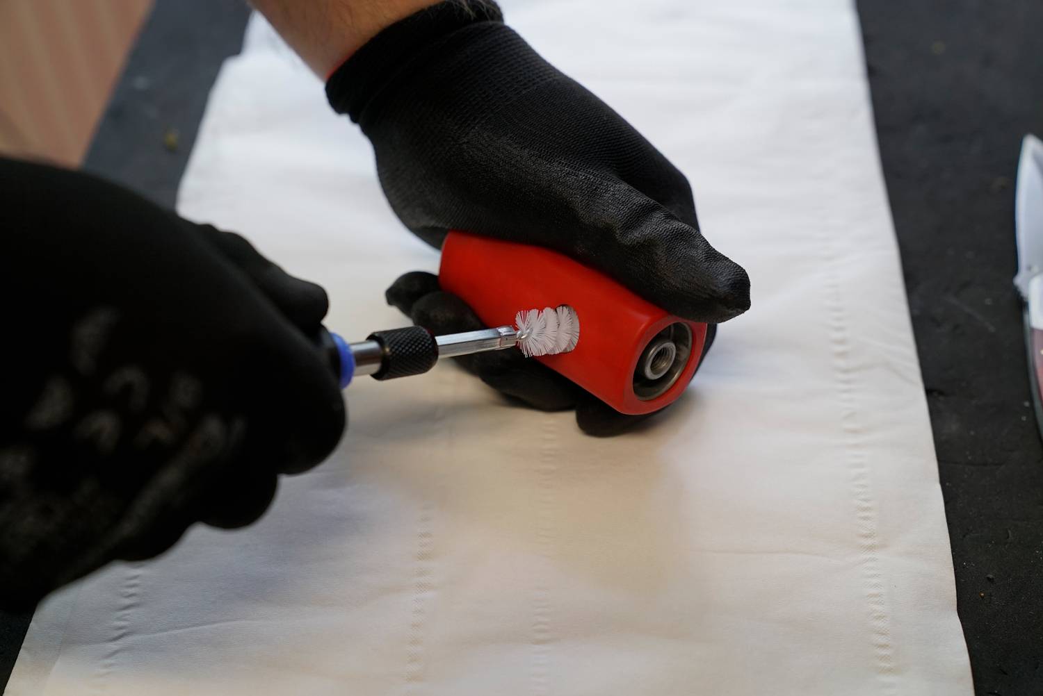 Een persoon draagt zwarte handschoenen en reinigt een rood kogellager van een skateboard-wiel met een borstel.