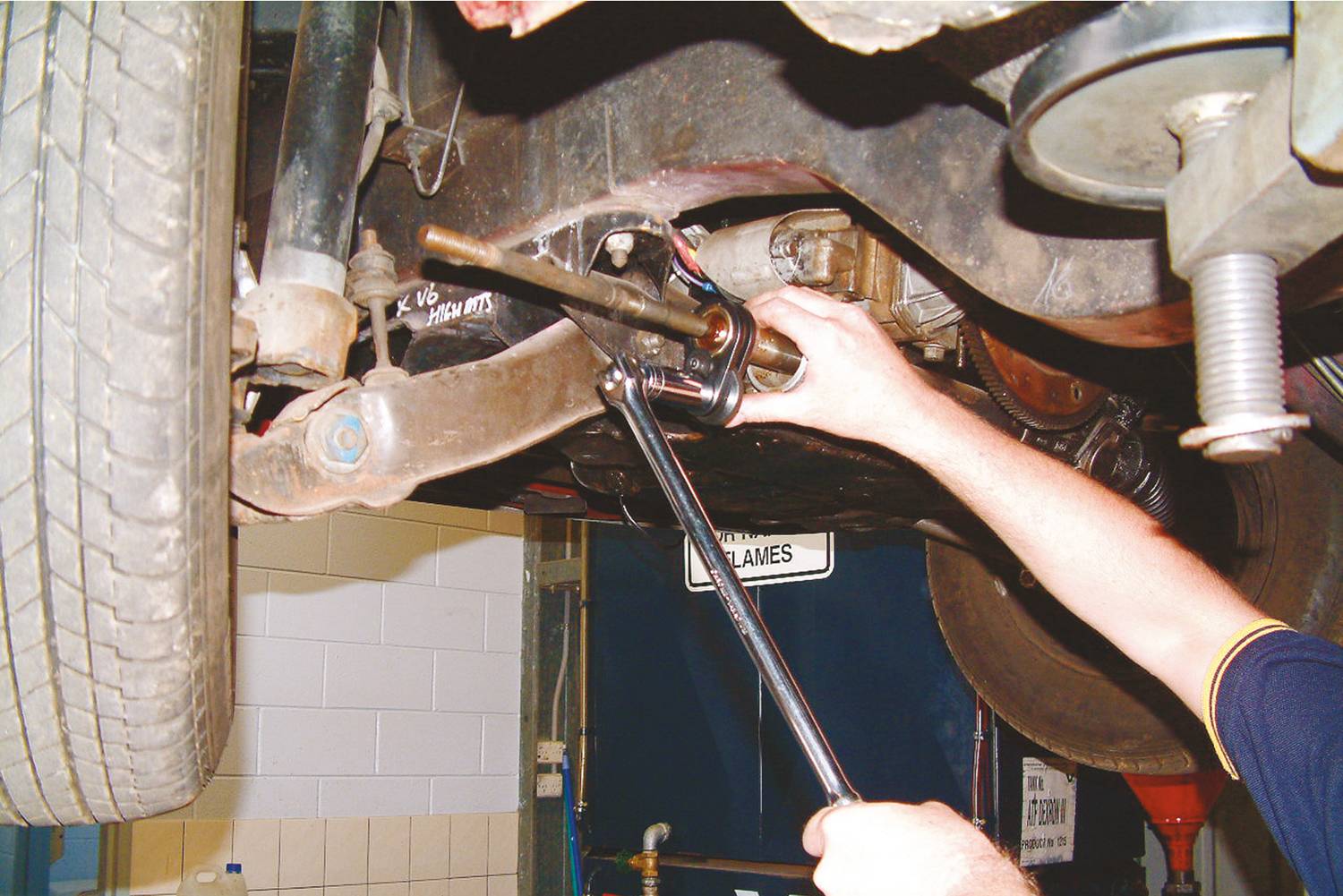 Een persoon repareert een auto van onderaf in een werkplaats. Ze gebruikt een moersleutel om aan een voertuigas of een onderstelonderdeel te werken.