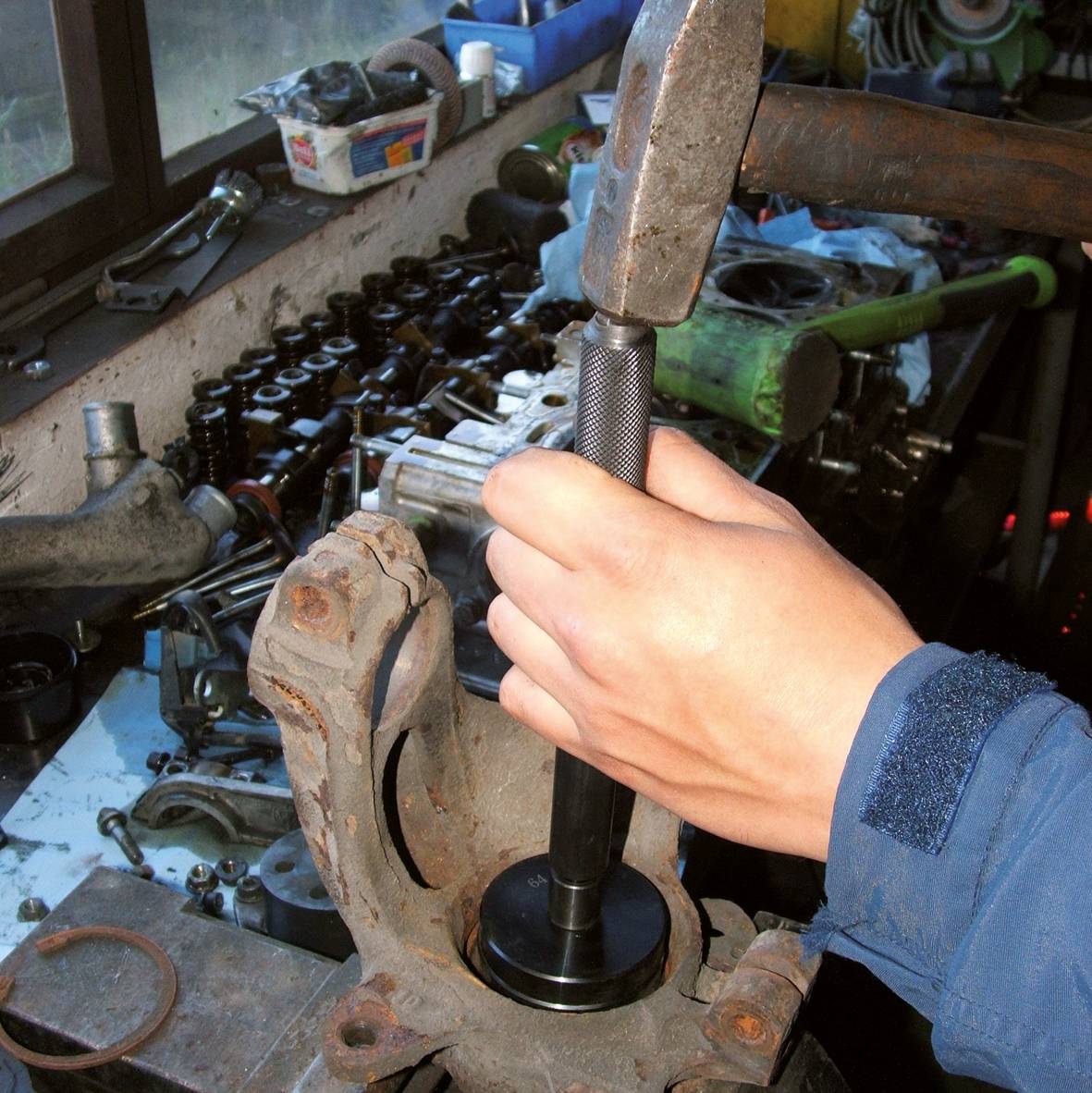 Een persoon gebruikt een gereedschap om een lager in een metalen beugel te plaatsen. Op de achtergrond zijn meer auto-onderdelen zichtbaar.