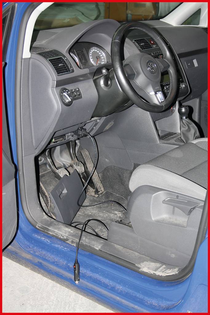 Een open auto met een blauwe portierlijst toont het interieur en de pedalen. Een kabel is zichtbaar op de vloer, die uit het dashboard komt.