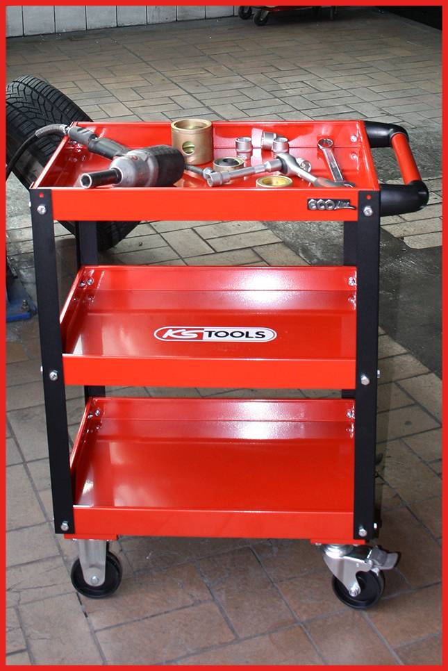 Rode werkplaatswagen met drie opbergvlakken, waarop verschillende gereedschappen zoals een boormachine, in een autogarage.