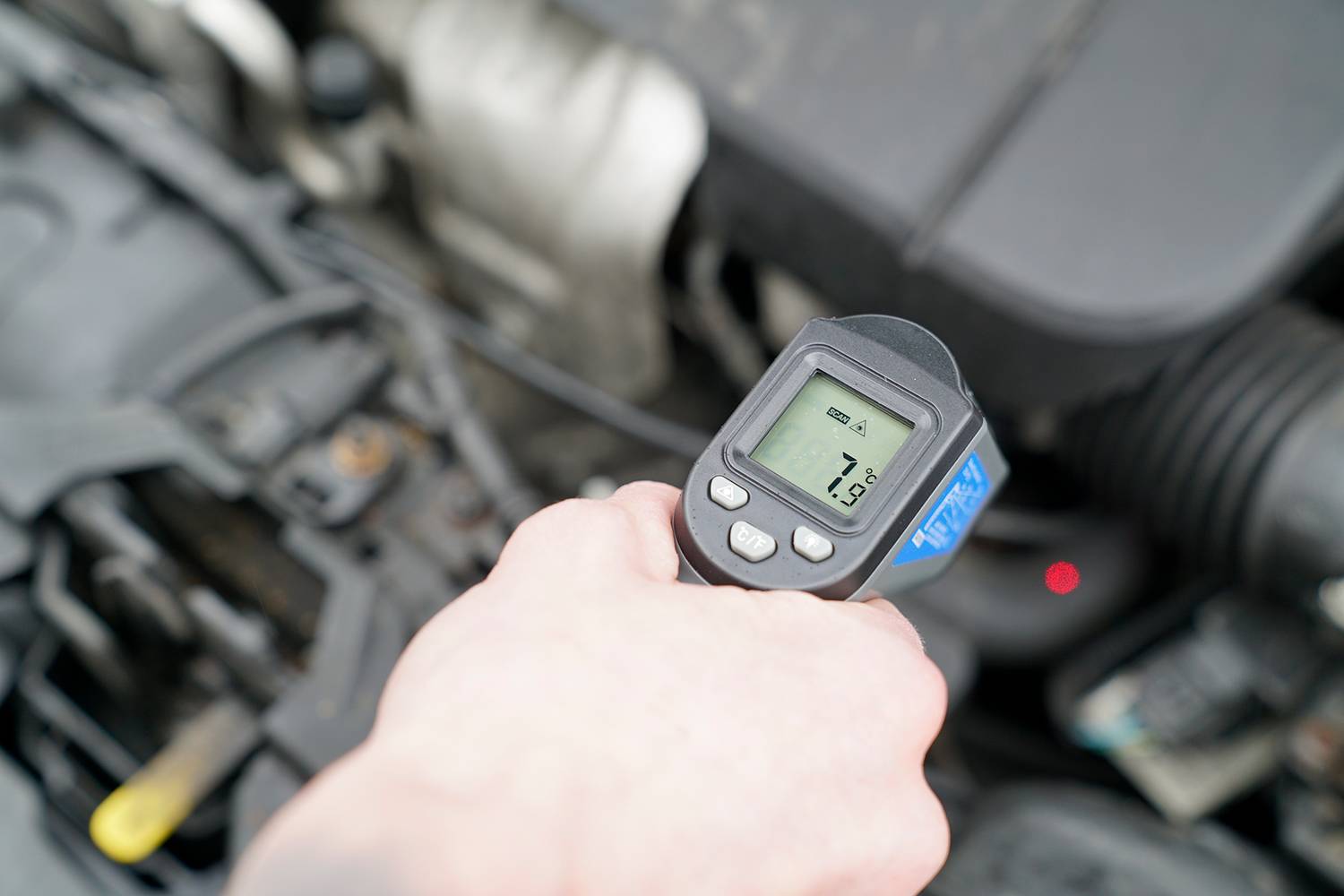 Een hand houdt een infrarood-thermometer boven een geopende automotor en toont een temperatuur van 51 graden Celsius aan.