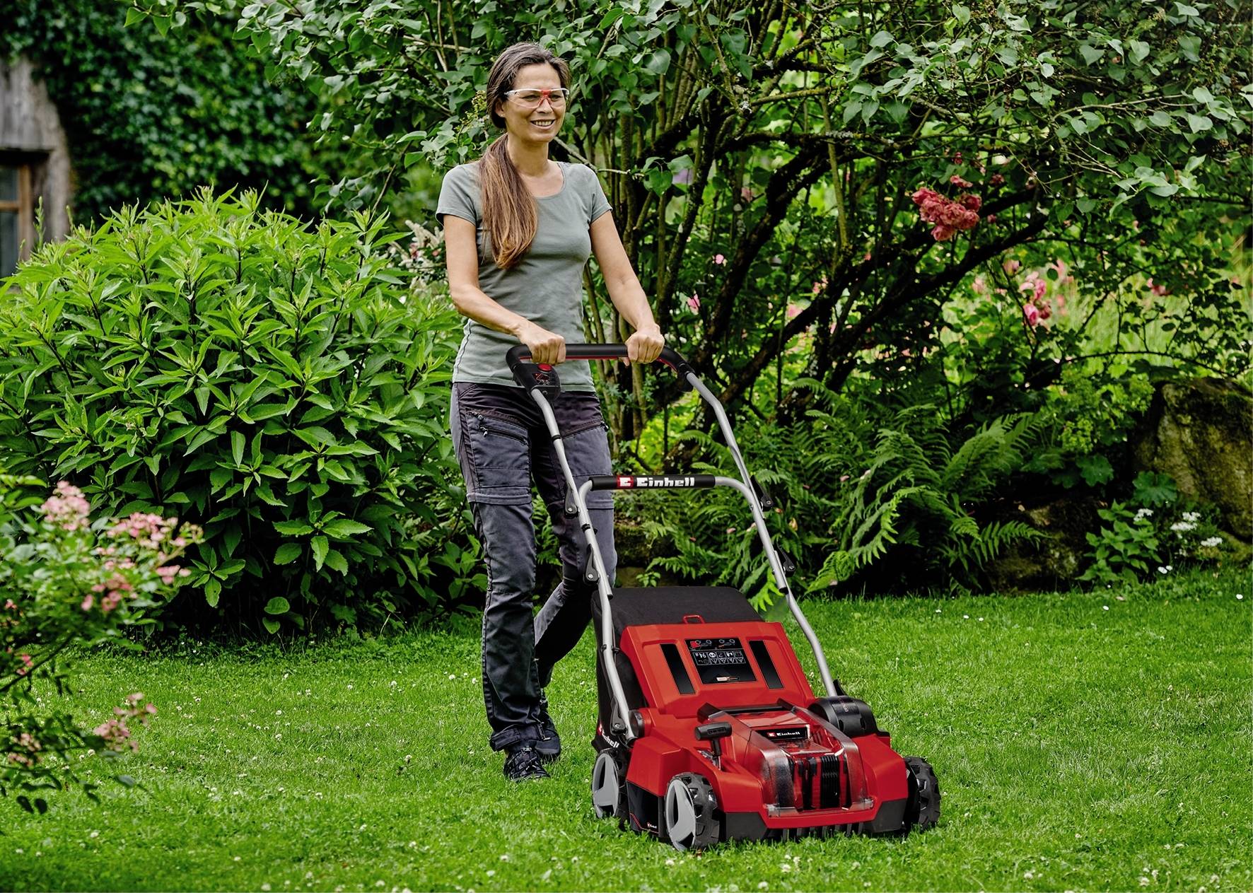 Een vrouw maait het gazon met een rode grasmaaier in een groene tuin. Ze draagt een grijze broek en een groen T-shirt.