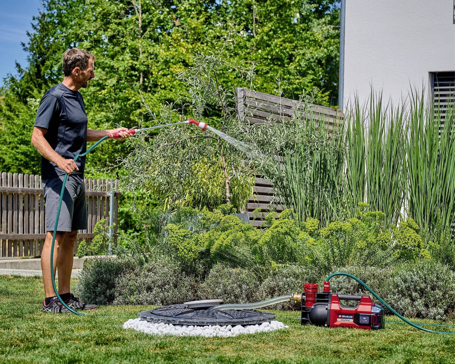 Een man besproeit planten in de tuin met een tuinslang. Een rode waterpomp ligt op het gazon in de buurt.