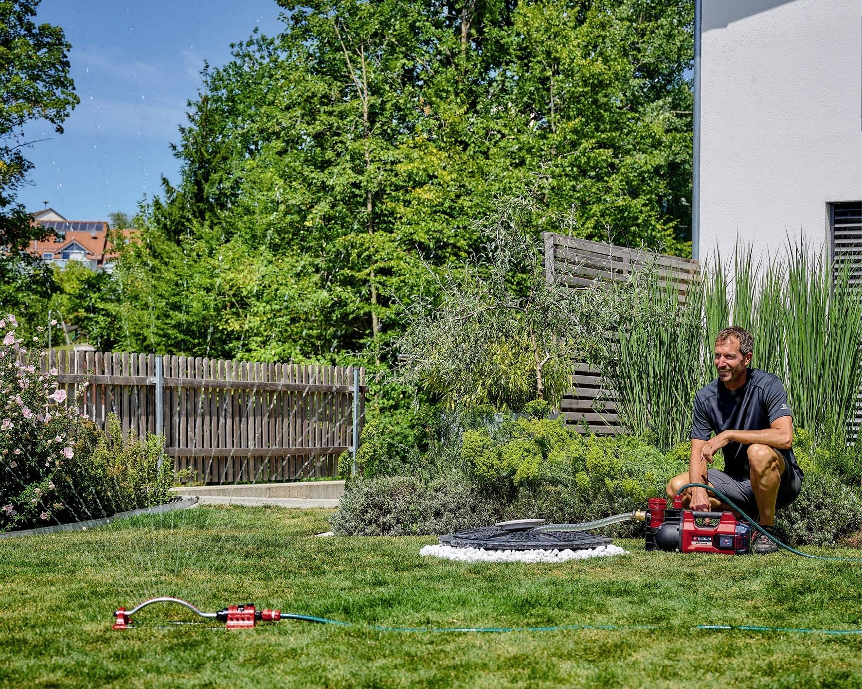 Een man knielt in de tuin naast een sproeier die het gazon besproeit. Op de achtergrond zijn bomen en een huis te zien.