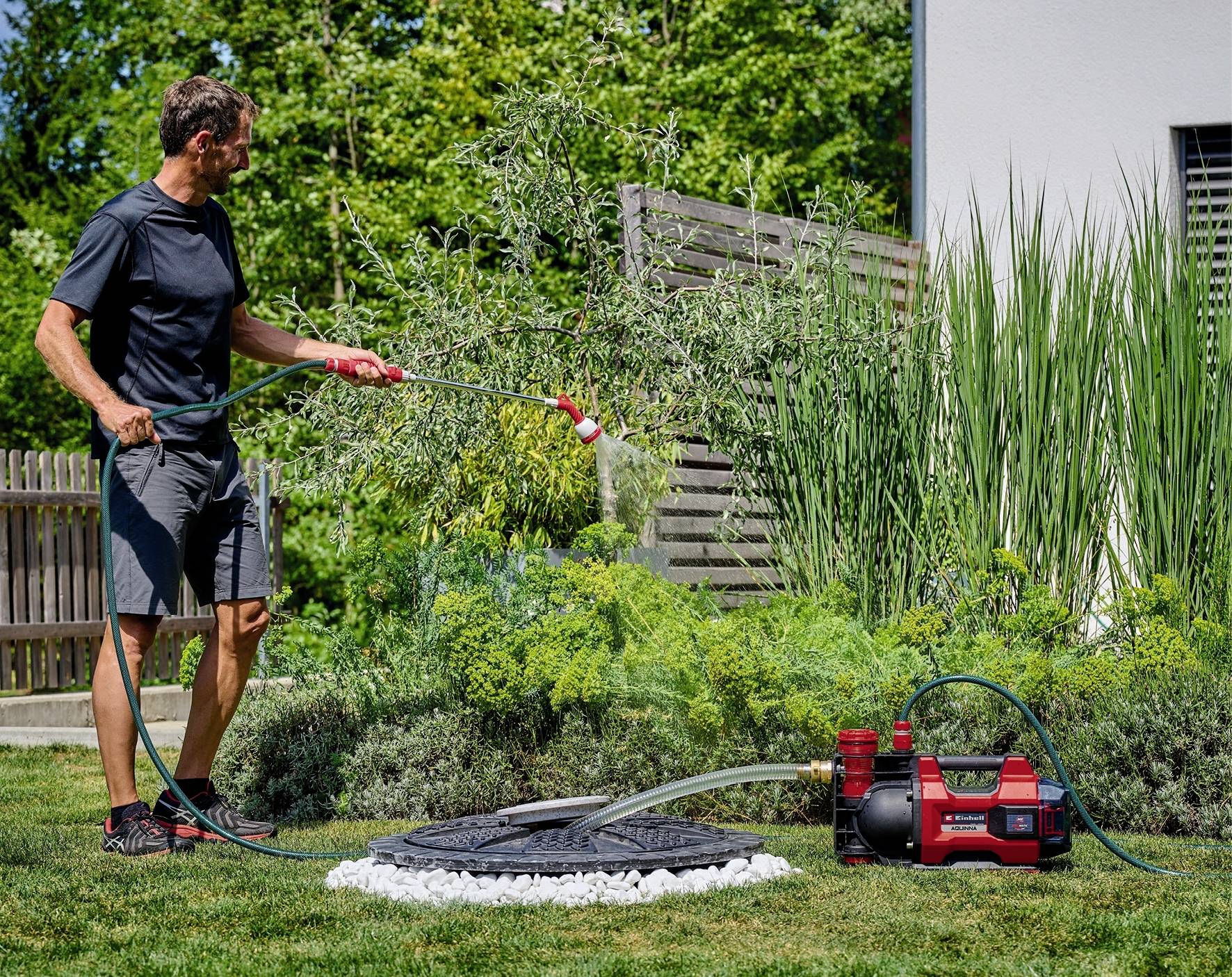 Een man besproeit planten met een tuinslang in een tuin. Naast hem staat een rode waterpomp. Op de achtergrond zijn bomen.