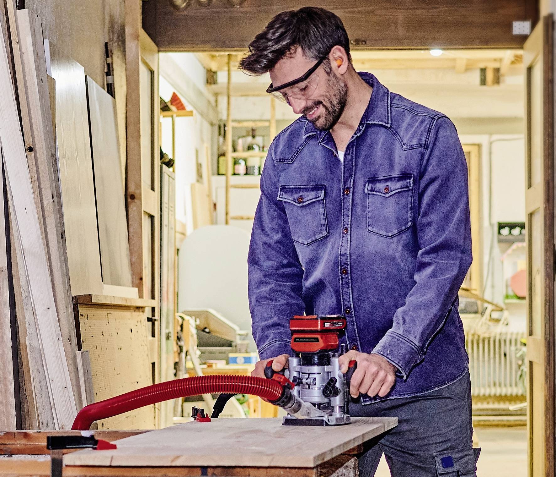 Een man in werkkleding gebruikt een houtfreesmachine in een werkplaats. Met een veiligheidsbril draagt hij, concentreert hij zich op de houten plaat.