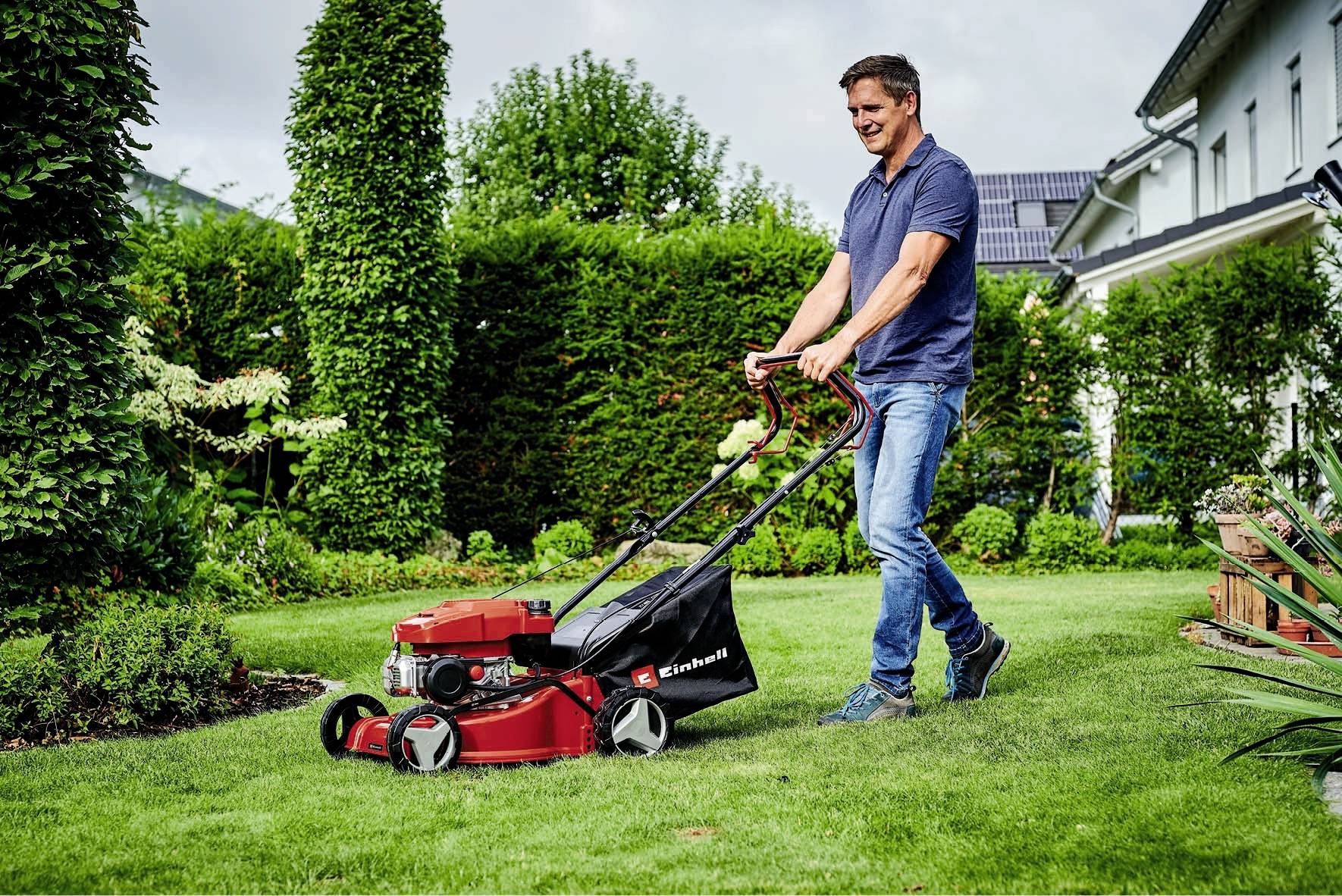 Een man maait het gras in een tuin met een rode grasmaaier. Op de achtergrond zijn struiken, bomen en een huis zichtbaar.