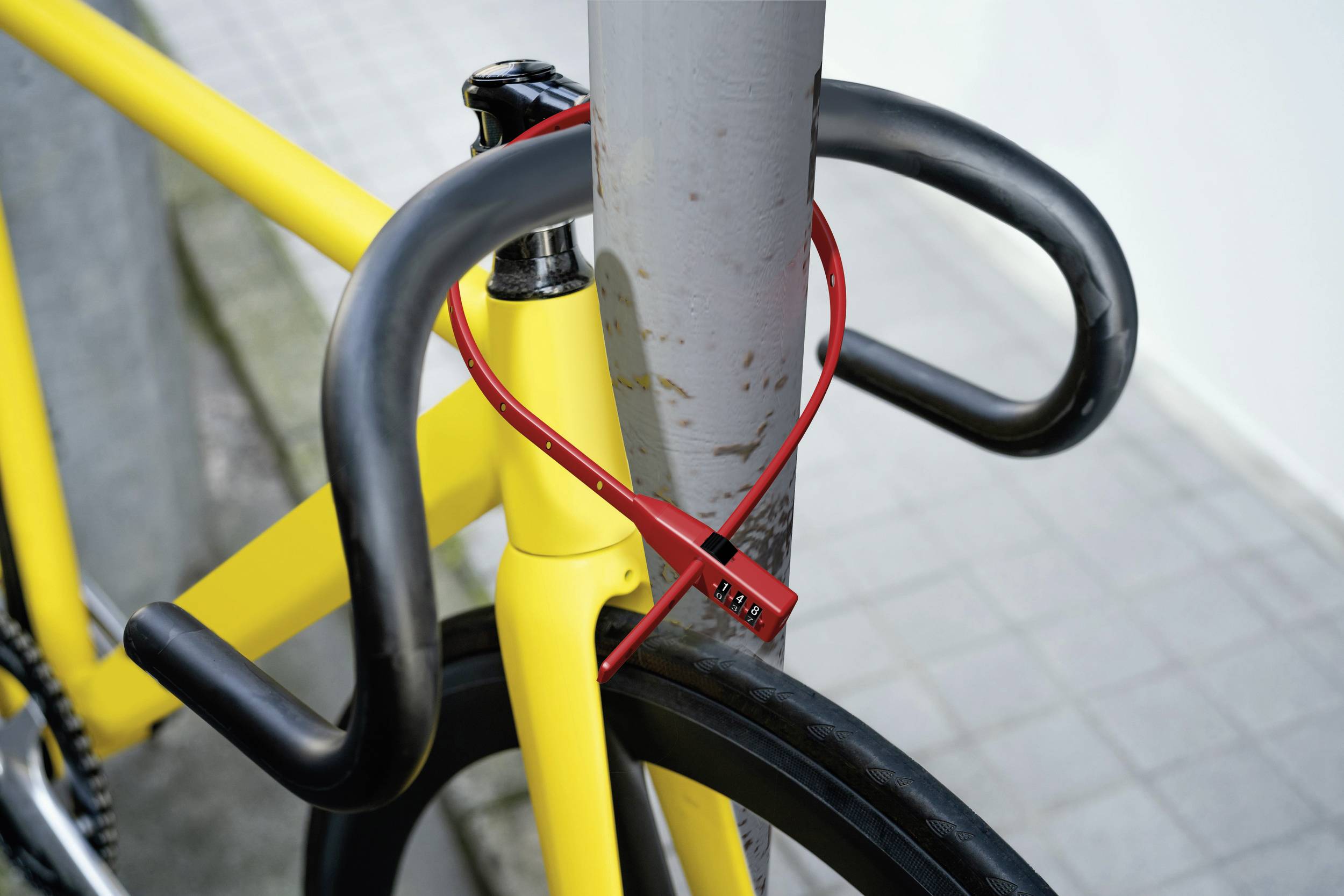 Een gele fiets is met een rood slot bevestigd aan een grijze metalen paal. De achtergrond toont stoeptegels.