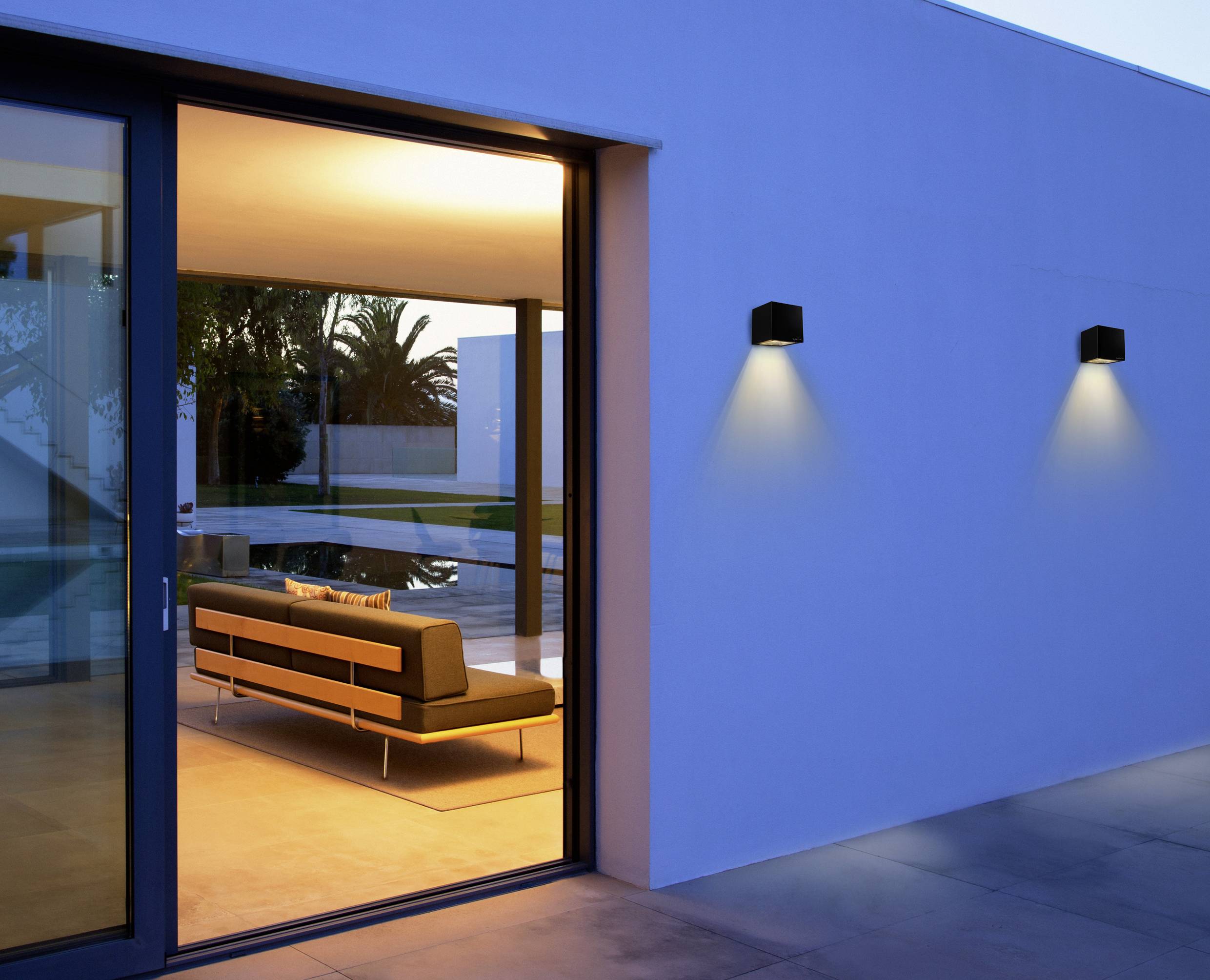 Een moderne lounge met verlichte binnenkant en een groot raam dat uitzicht biedt op een terras met zwembad en palmbomen.