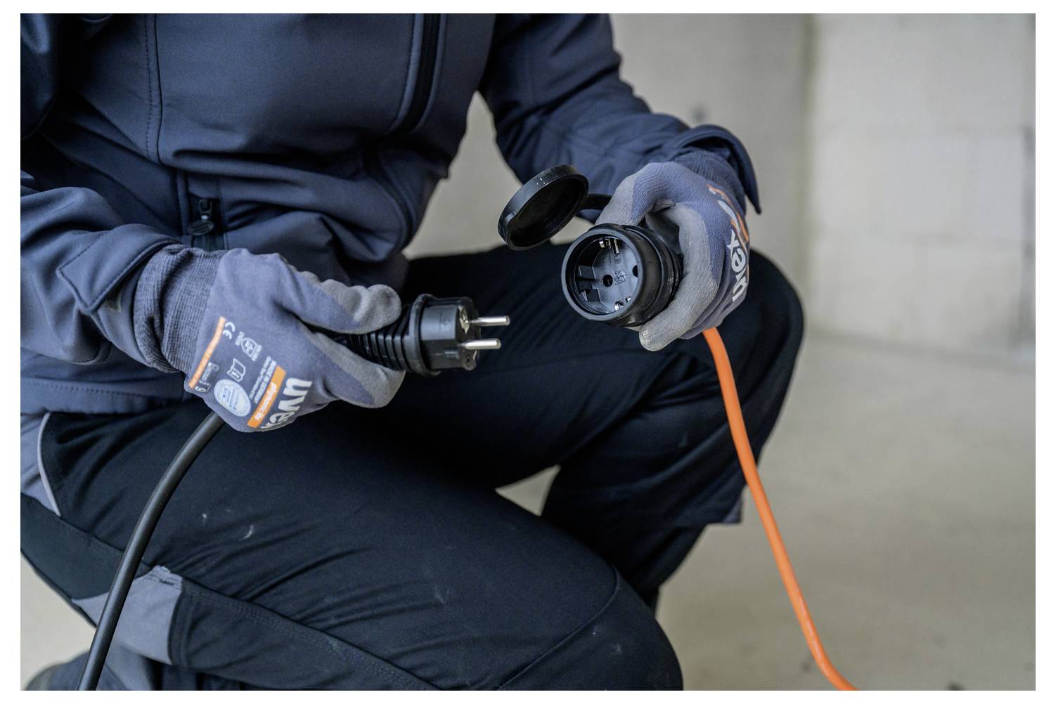 Persoon met handschoenen steekt een oranje verlengsnoer in een zwarte stroomconnector, ter voorbereiding van een elektrische verbinding.
