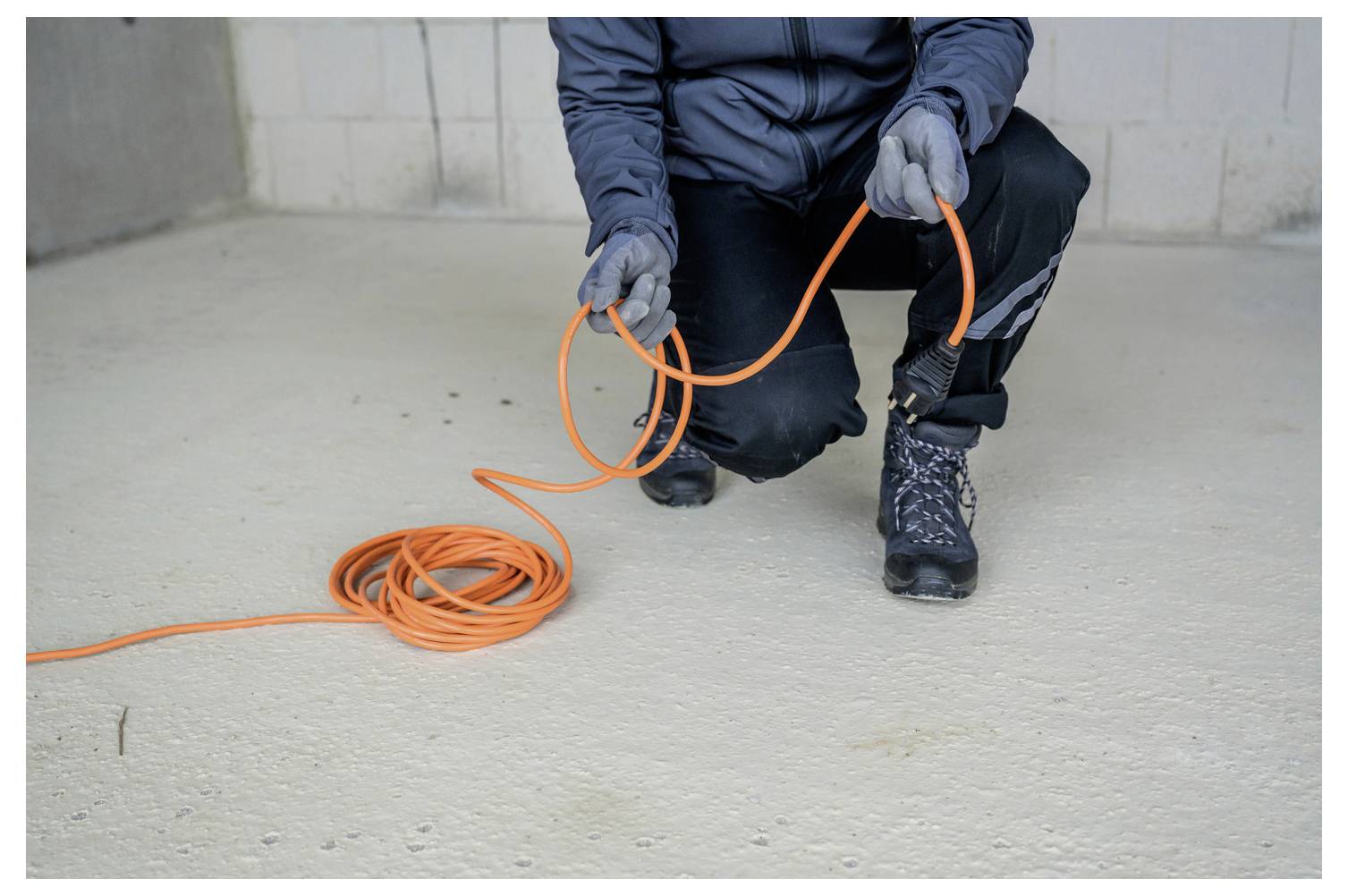 Arbeider in handschoenen knielt op een betonnen vloer en houdt een felOranje kabel of verlengkabel vast terwijl hij deze uitrolt.